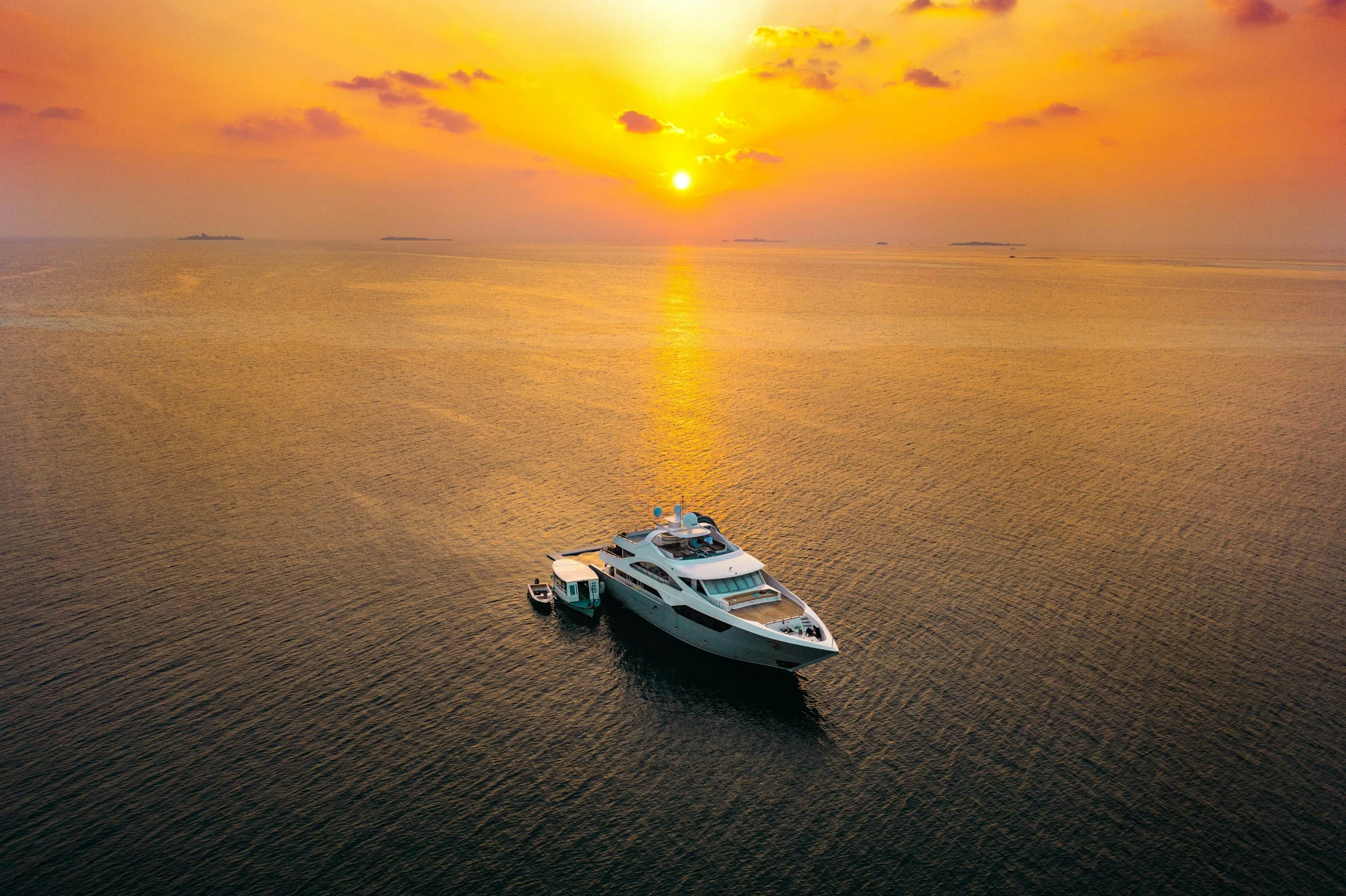 A white yacht anchored in calm water during a vibrant sunset, with the sky filled with orange, yellow, and pink clouds.