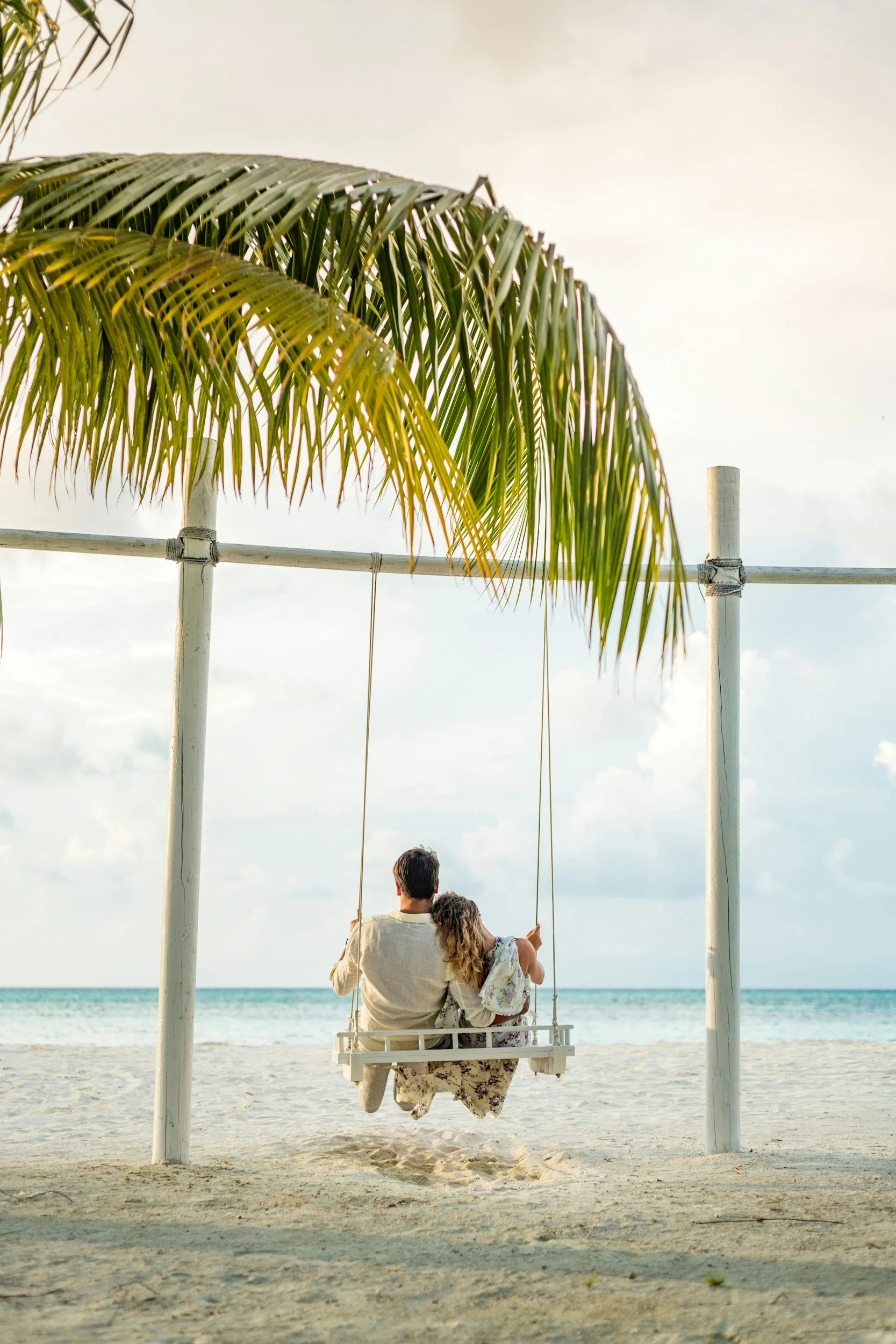 A couple sitting on a swing at the beach, with palm trees overhead and the ocean in the background.