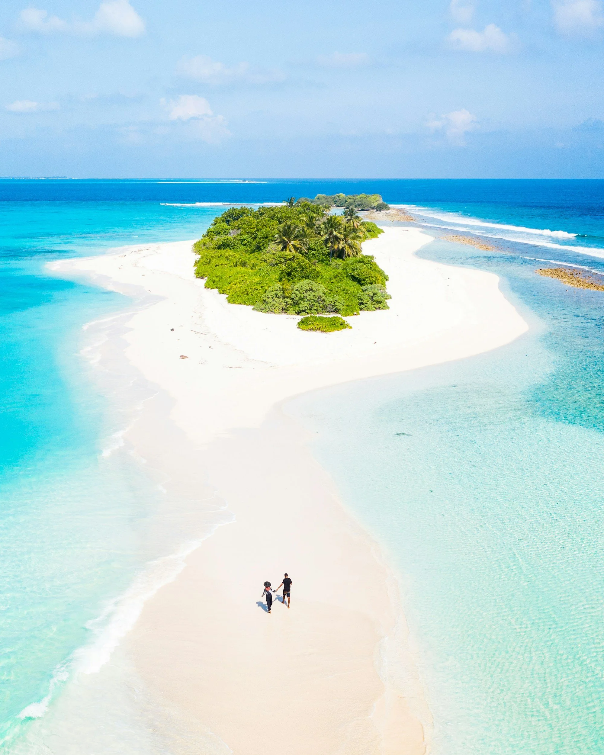 An aerial view of a tropical island with white sandy beaches, lush green trees, and turquoise waters surrounding it, with two people walking on the beach.