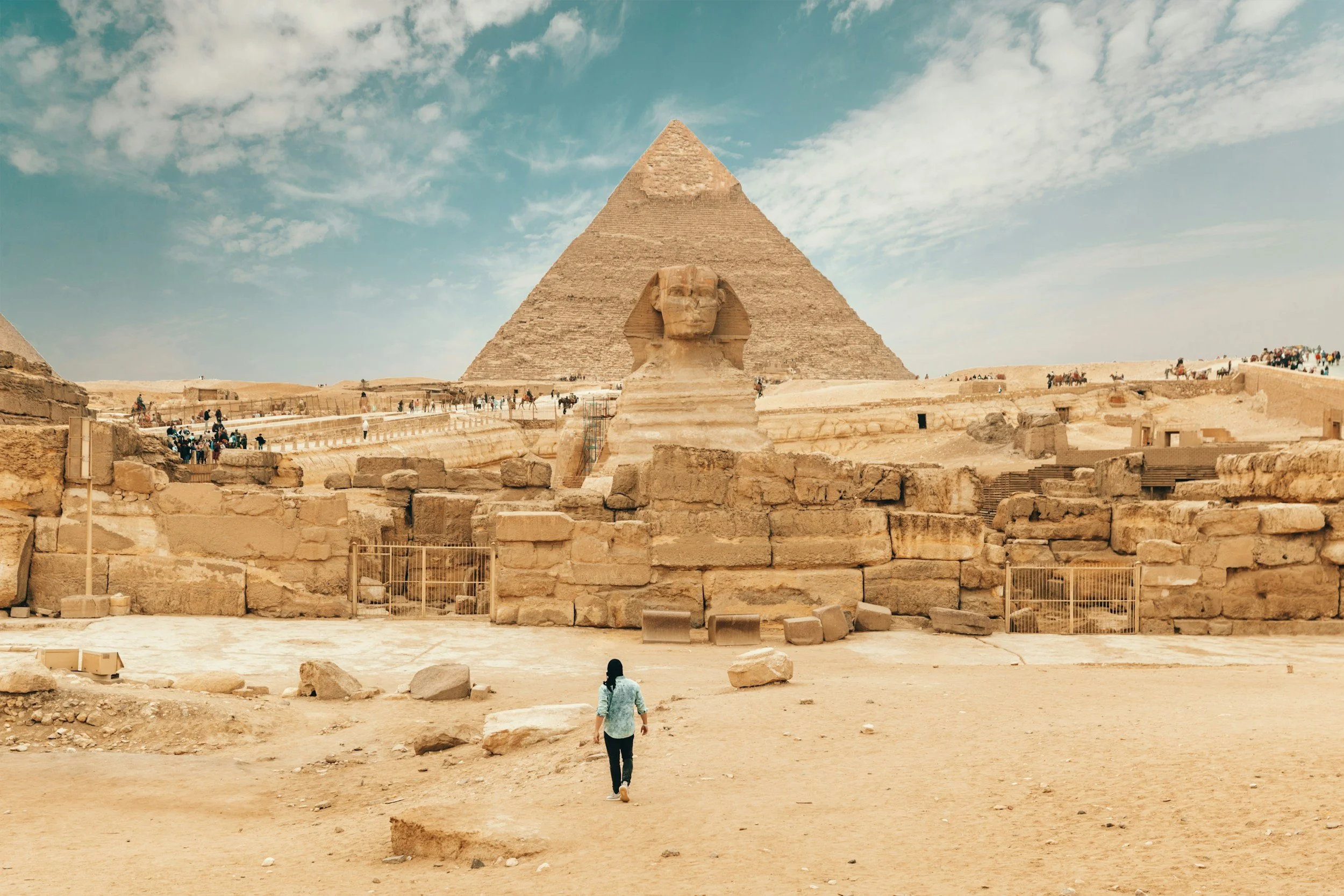 A person walking towards the Great Pyramid of Giza with the Great Sphinx of Giza in the foreground, under a partly cloudy sky.