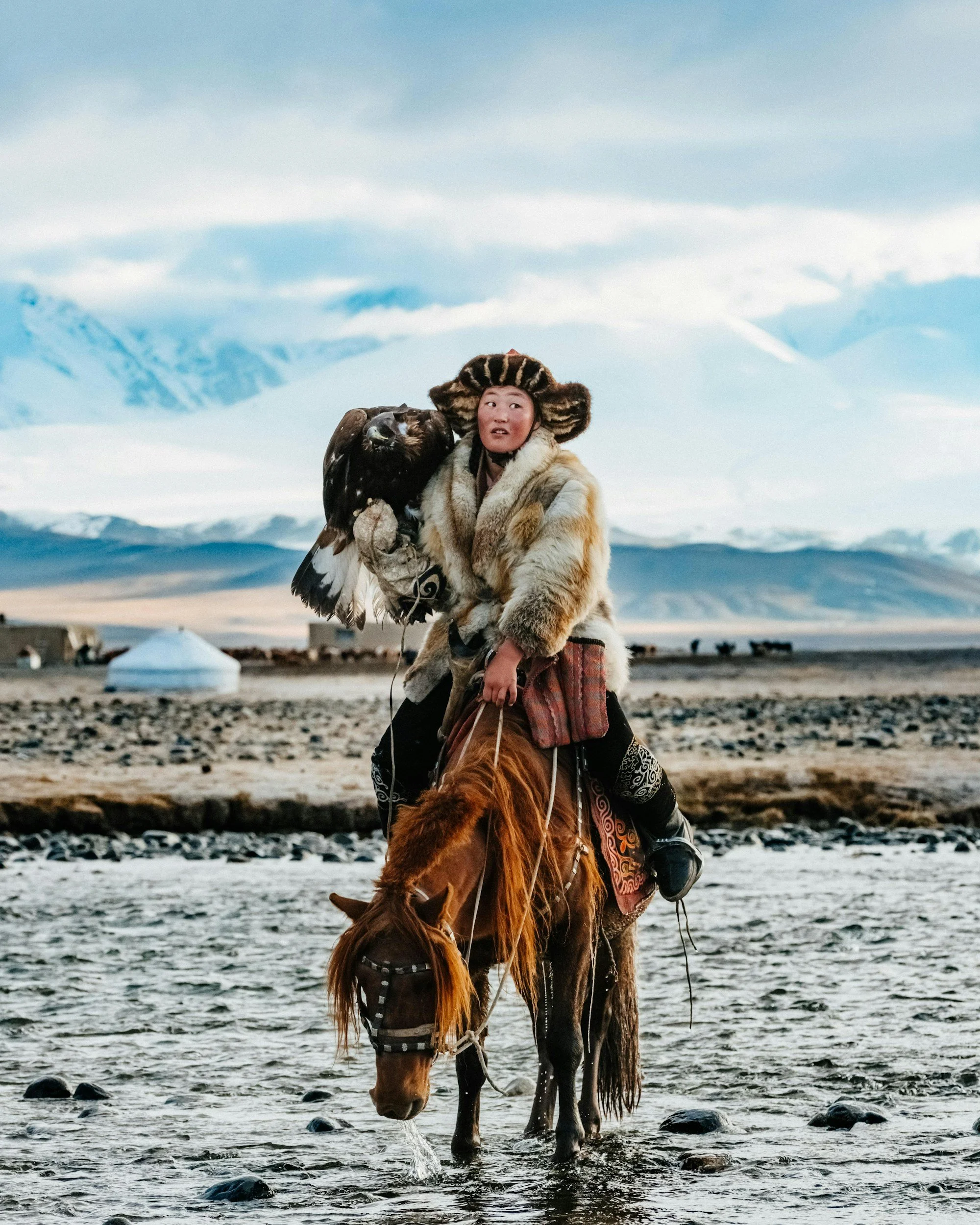 Person wearing traditional fur clothing riding a horse through a shallow stream, holding a bird of prey, with mountains and a blue sky in the background.