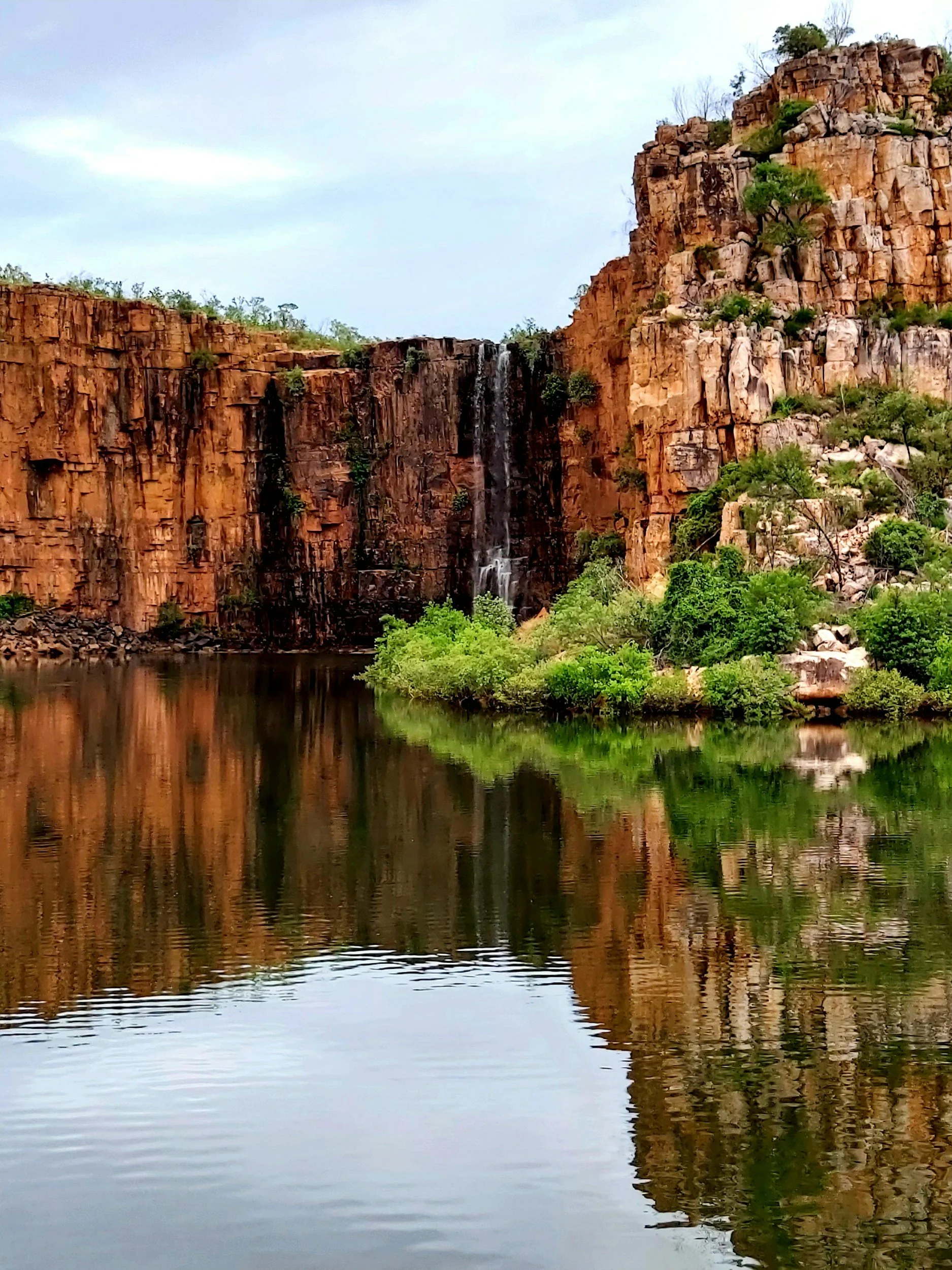 Scenic view of red rock cliffs with a waterfall, green bushes, and a calm body of water reflecting the landscape.
