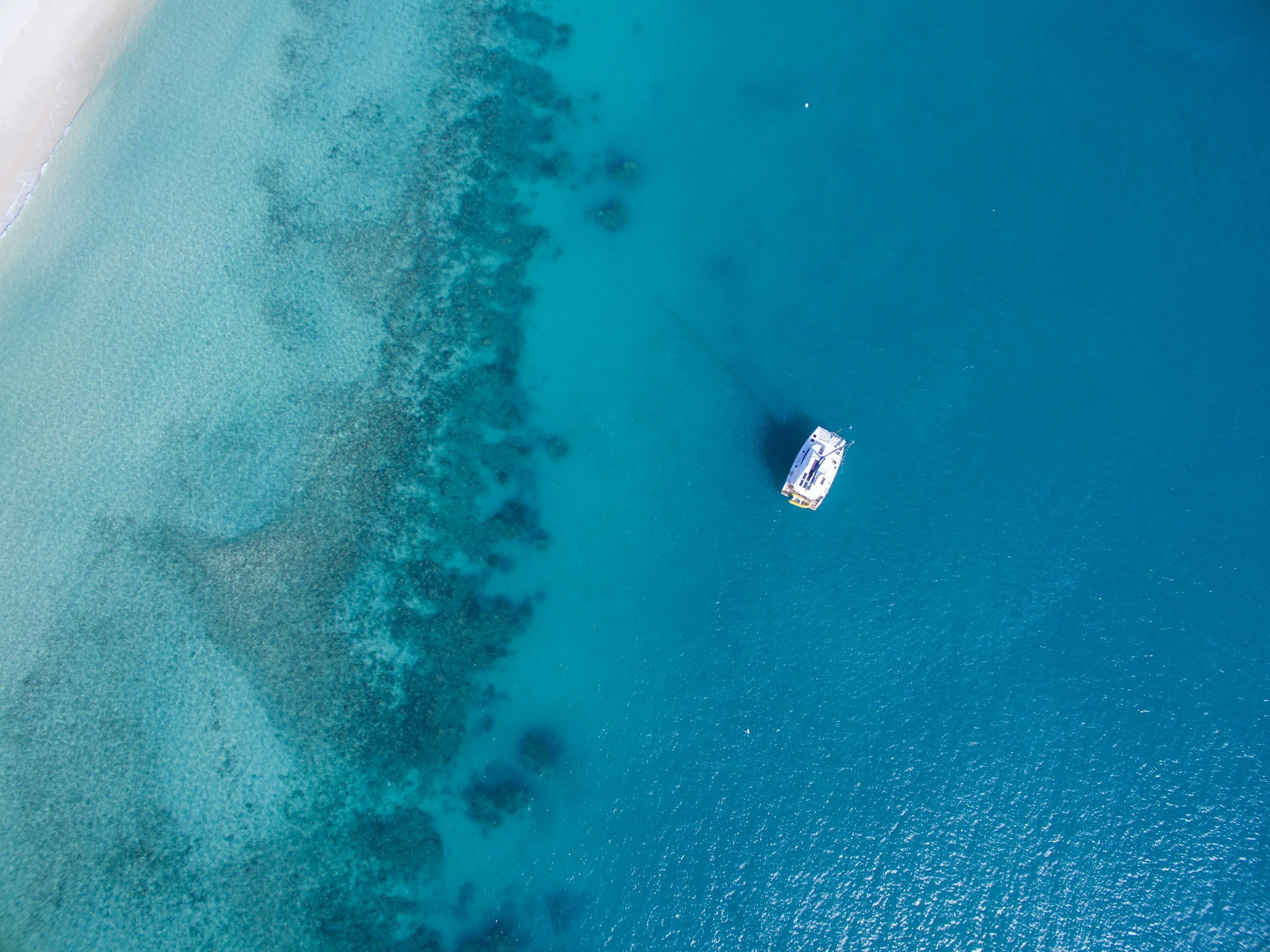 An aerial view of a boat anchored in clear turquoise water near a sandy shoreline with dark patches of underwater vegetation.