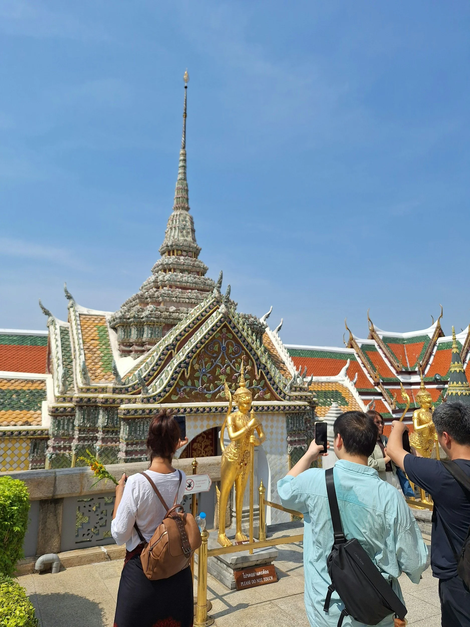 Tourists taking photos of a golden statue outside a colorful Thai temple with traditional architecture and a tall spire under a clear blue sky.
