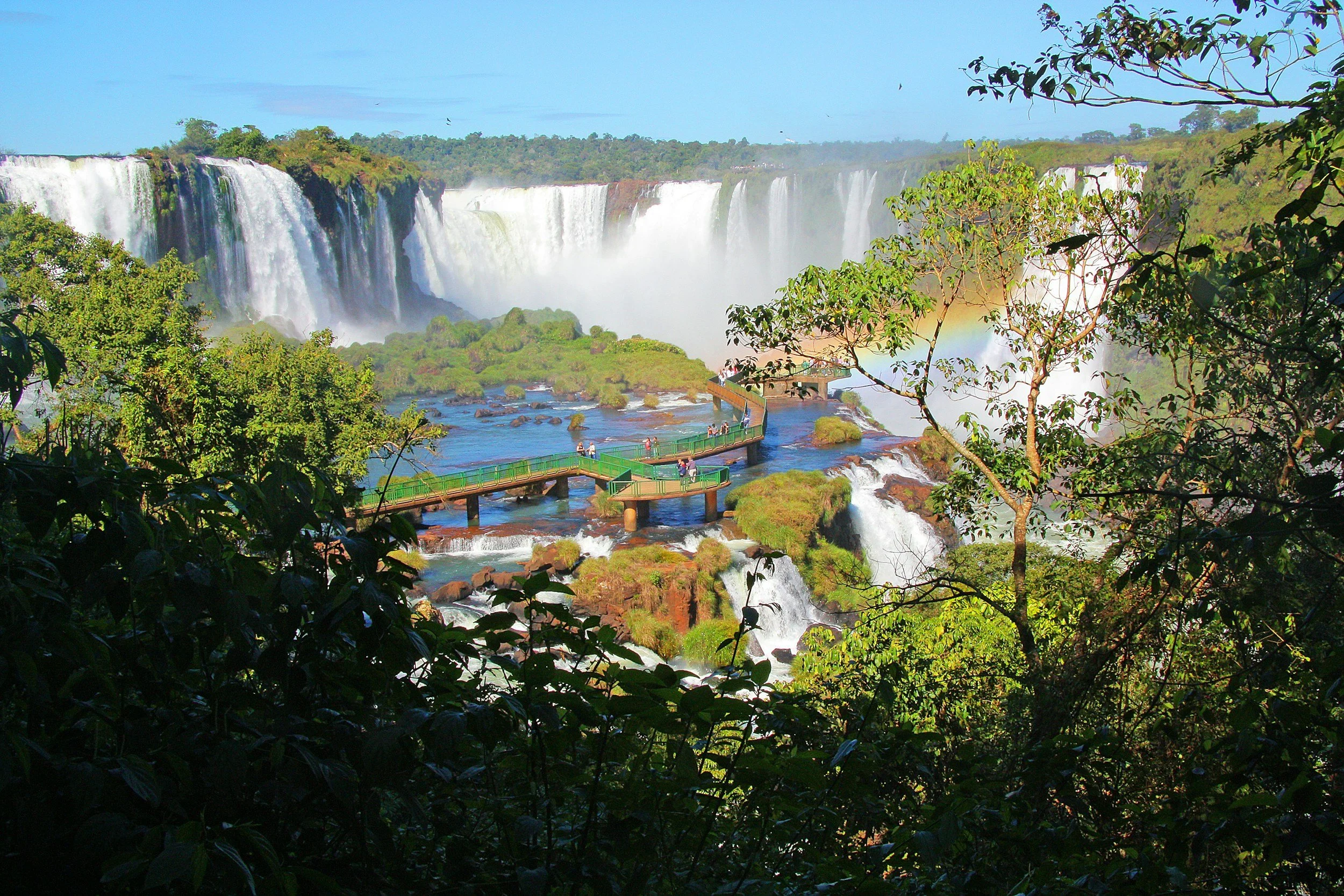 View of Iguazu Falls with cascading waterfalls, a rainbow, and a walkway with visitors among lush greenery.