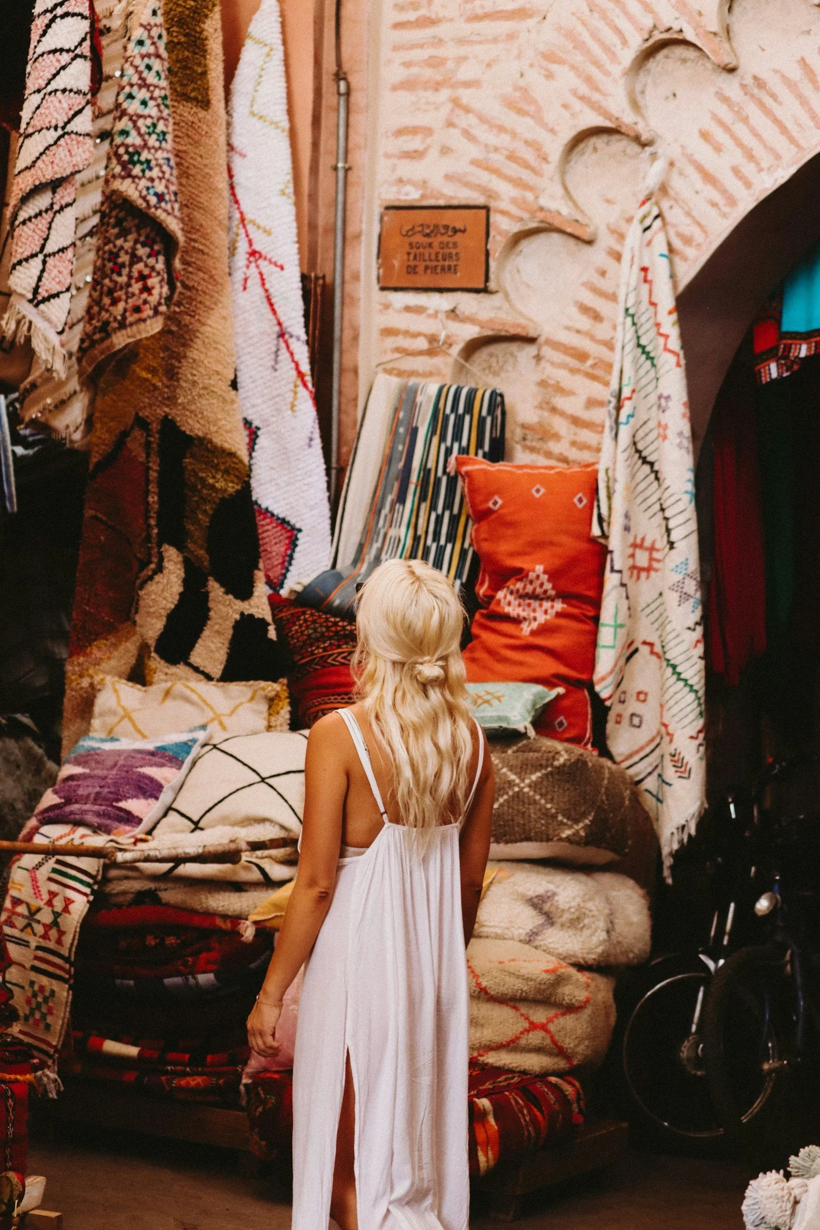 A woman with long blonde hair in a white dress looking at colorful rugs and cushions displayed for sale at a market or shop.