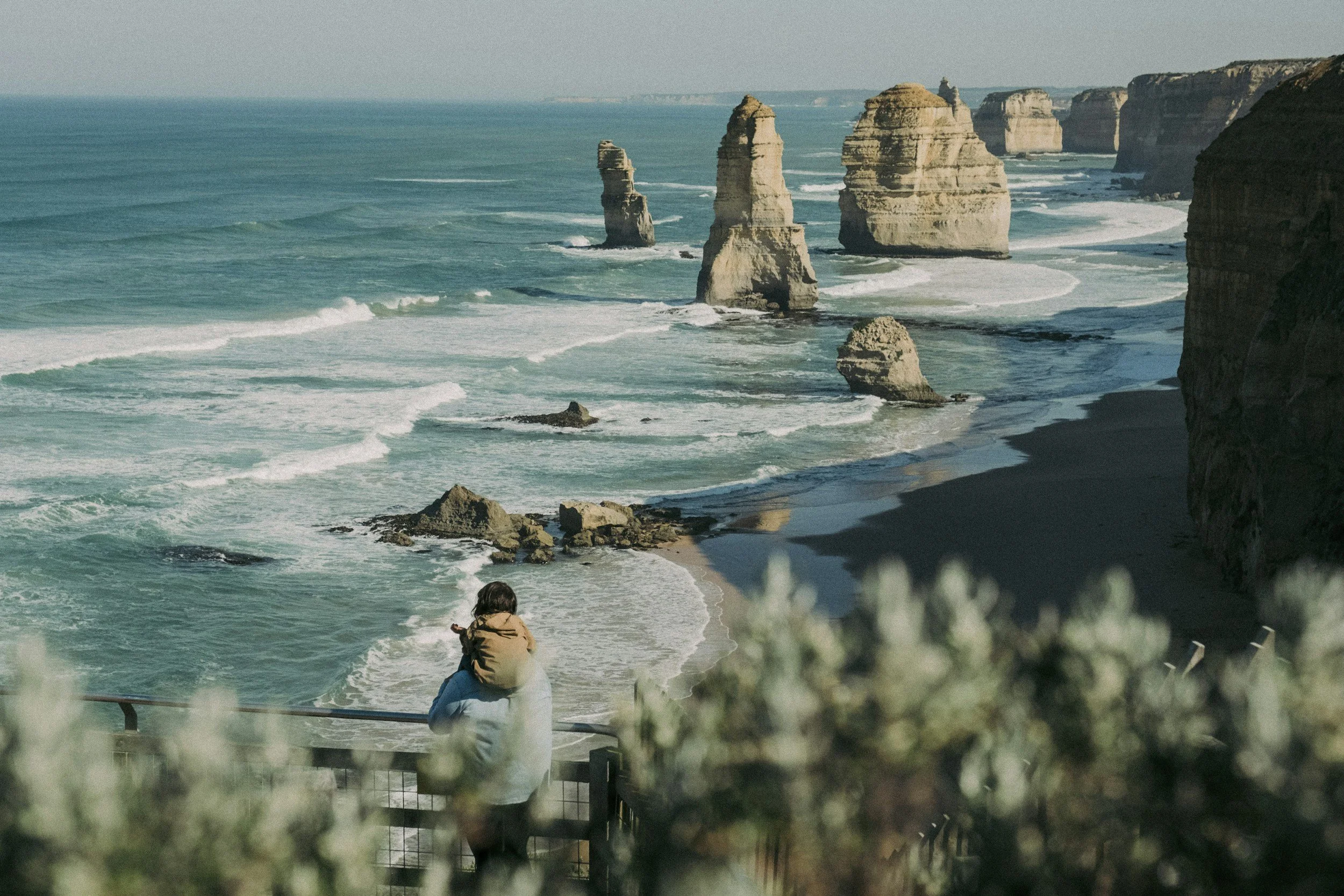 A person with a child on their shoulders standing at a lookout, looking at the ocean and rock formations called the Twelve Apostles along the coast of Australia.