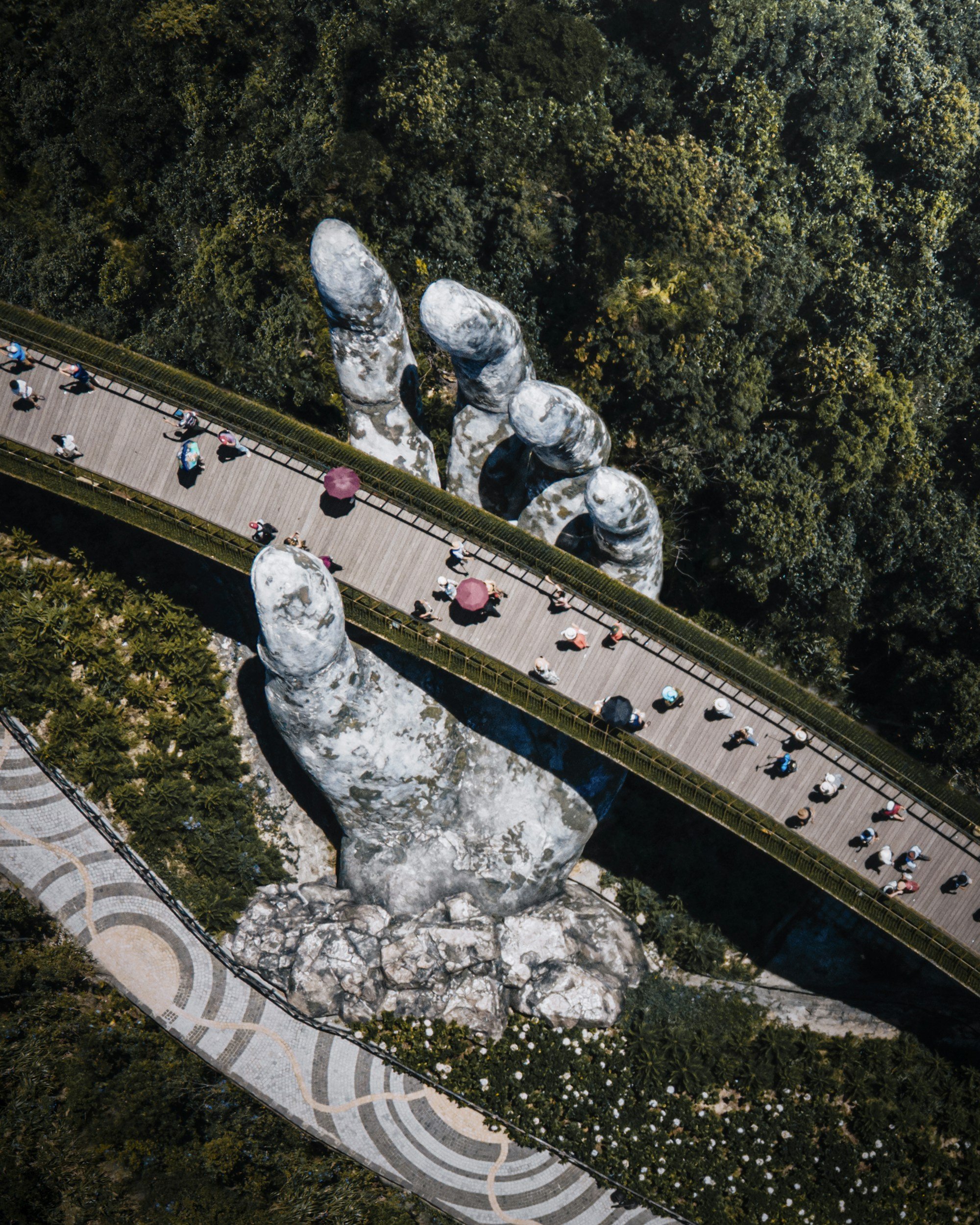 An aerial view of a large hand sculpture with four fingers, located in a park surrounded by greenery. A wooden walkway with people walking, some carrying umbrellas, runs through the scene.