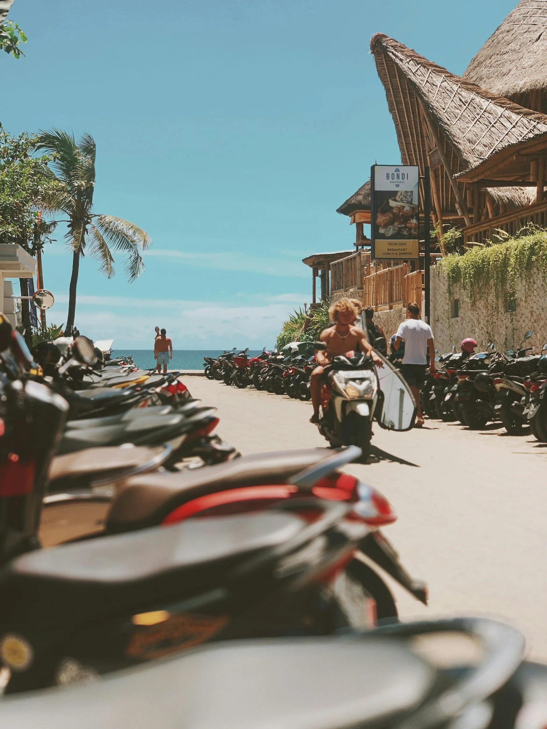 Cars parked along a street near the beach with a view of the ocean in the background. People are riding two scooters, and there are tropical trees and thatched-roof buildings.