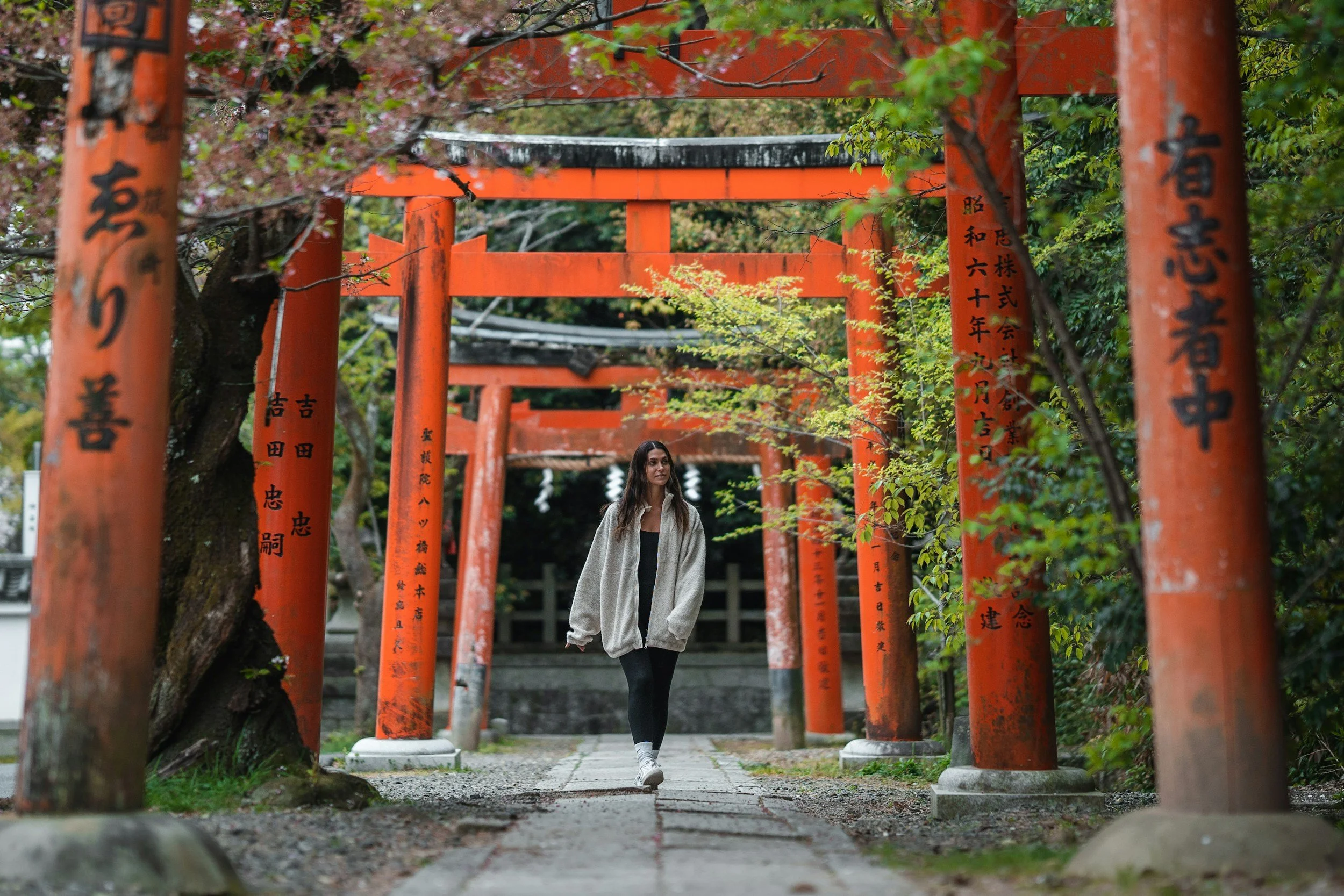 A woman walking through a pathway lined with orange Torii gates at a Japanese shrine, surrounded by green foliage.