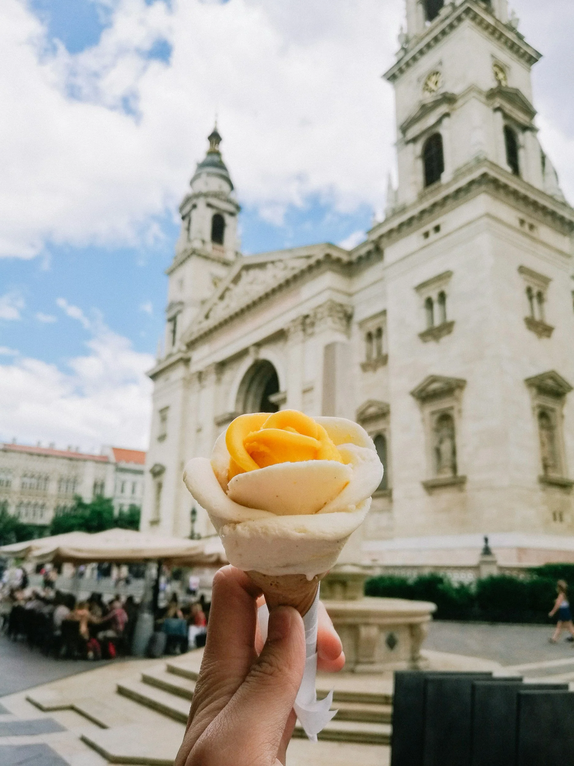 Hand holding ice cream cone with white and yellow swirl in front of a historic church with twin towers, steps, and outdoor seating.