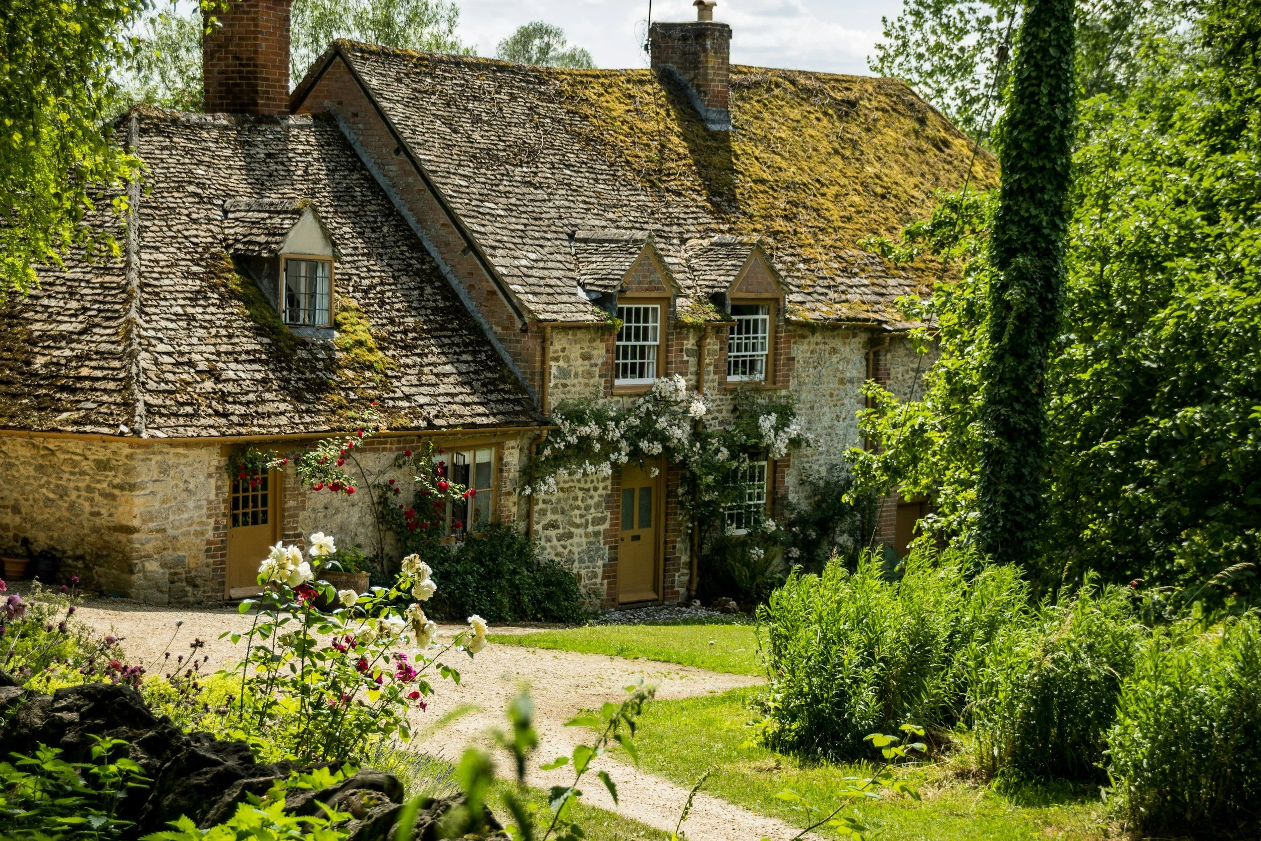 A rustic stone house with a moss-covered tiled roof, surrounded by lush greenery and blooming flowers.