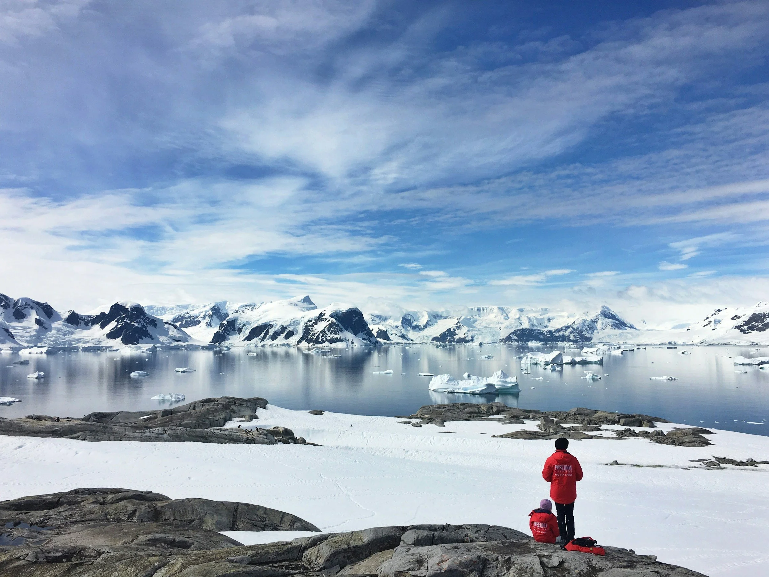 People dressed in red winter jackets sitting on rocks overlooking icy coastline with snow-covered mountains and floating icebergs under a blue sky with clouds.