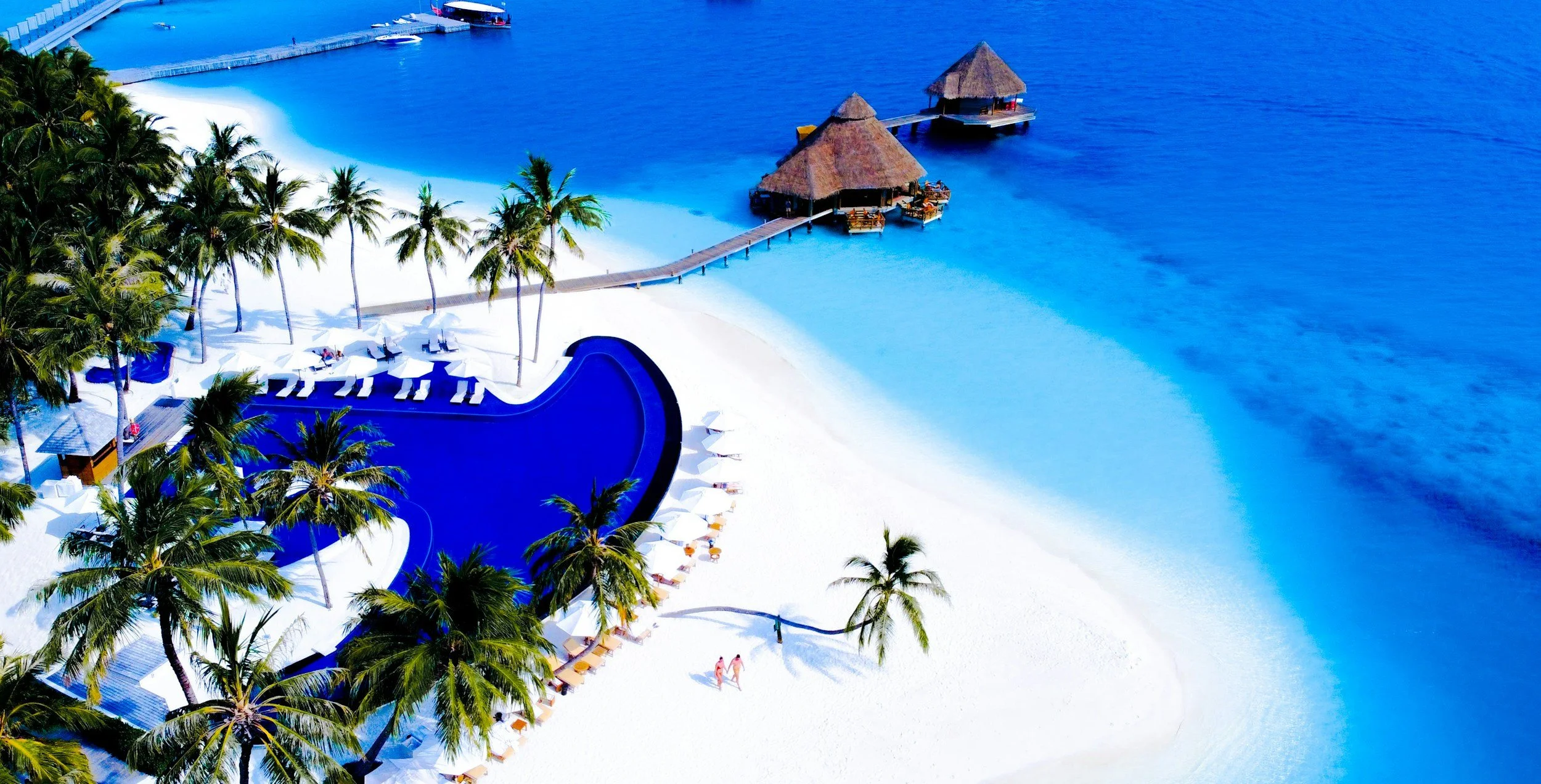 Aerial view of a tropical beach resort with a large blue pool, palm trees, white sand, overwater huts, and a pier extending into the ocean.
