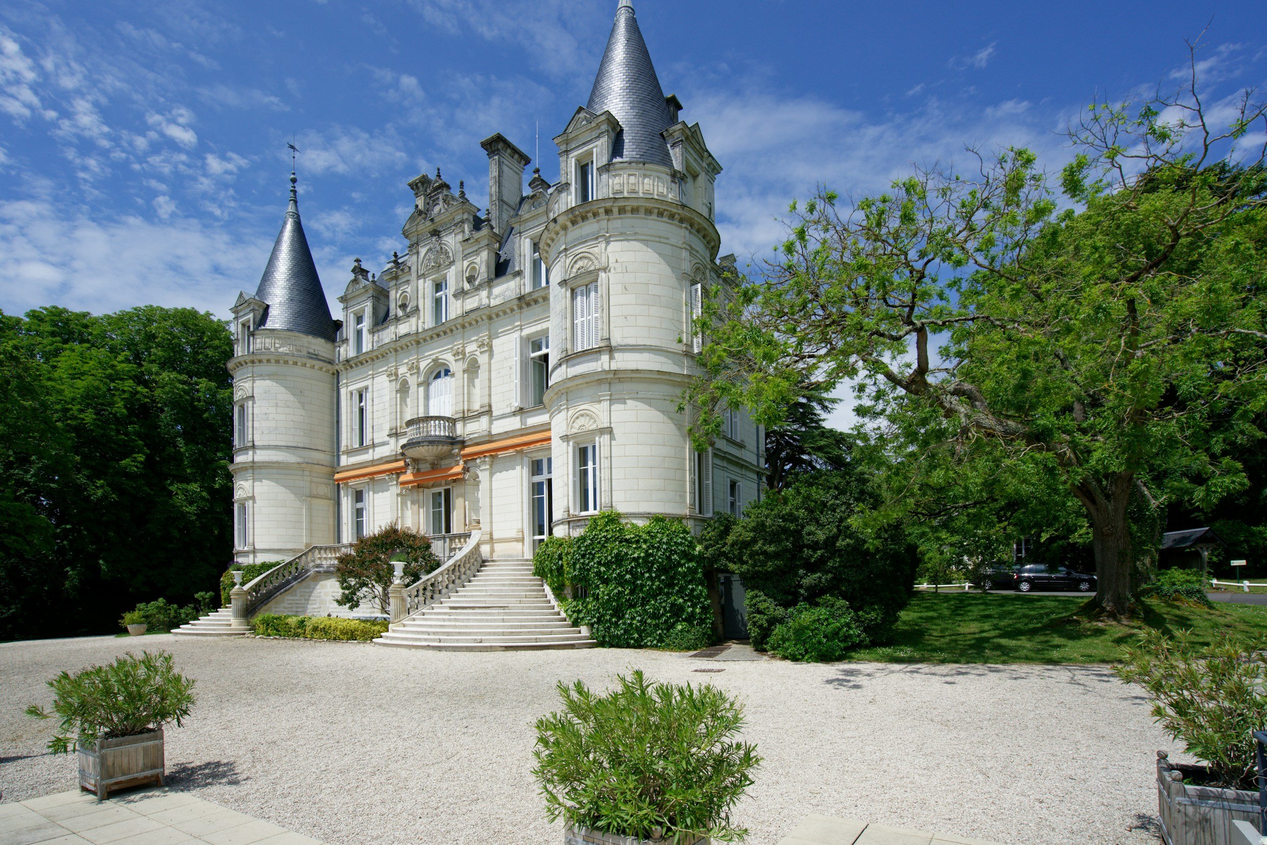 A large, white castle with multiple turrets and a steep, pointed roof, surrounded by lush green trees and a gravel courtyard under a partly cloudy blue sky.