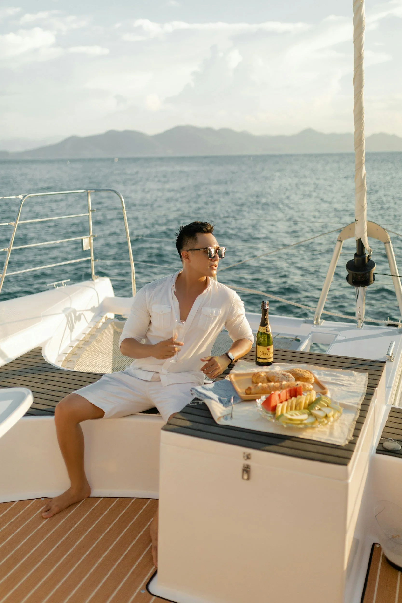 A man sitting on the deck of a yacht, holding a glass of champagne, with a tray of fruit and bread, and a bottle of champagne nearby, overlooking the ocean with mountains in the background.