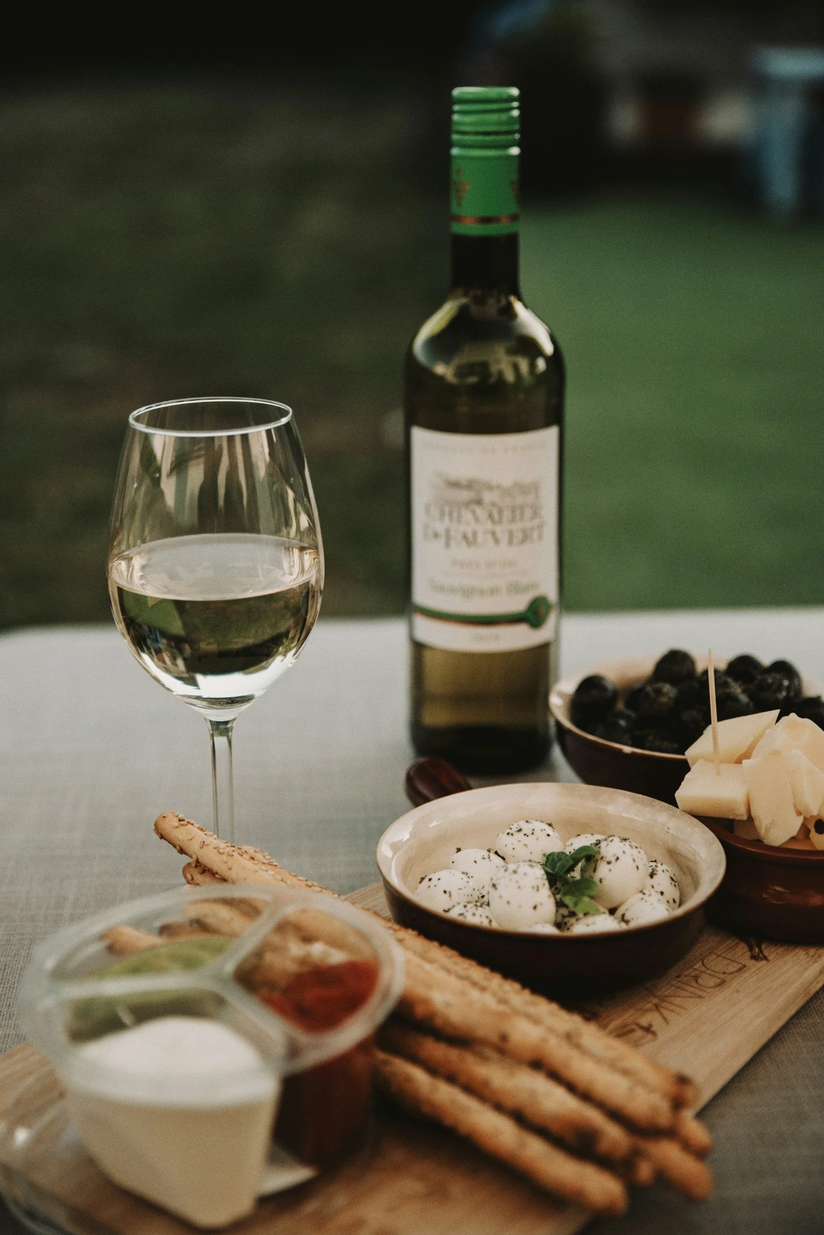 A wine glass with white wine, a bottle of Sauvignon Blanc, and various cheeses, olives, breadsticks, and condiments on a table outdoors.