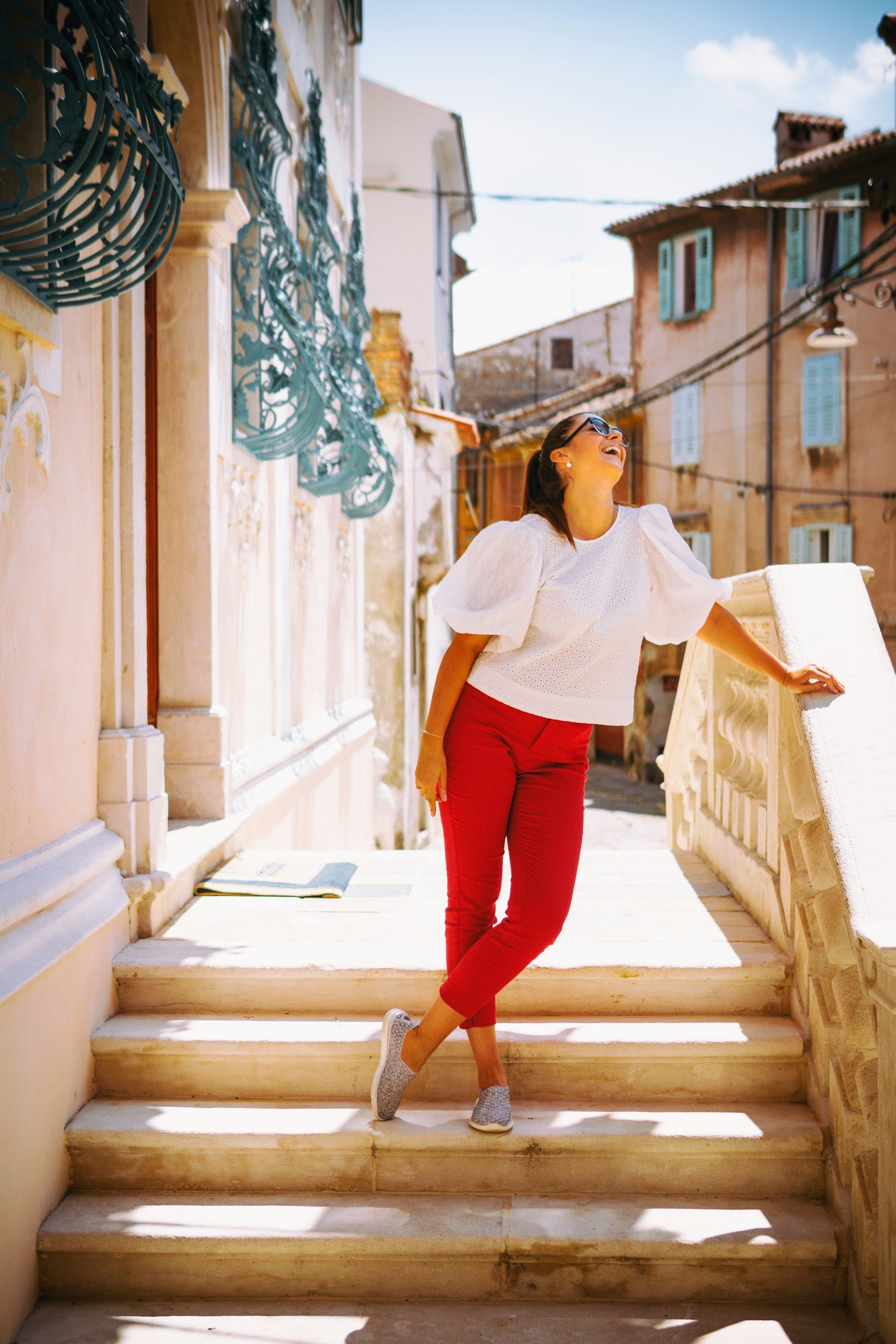 A woman with sunglasses laughing and leaning on a stone staircase railing in a sunny, colorful European town.