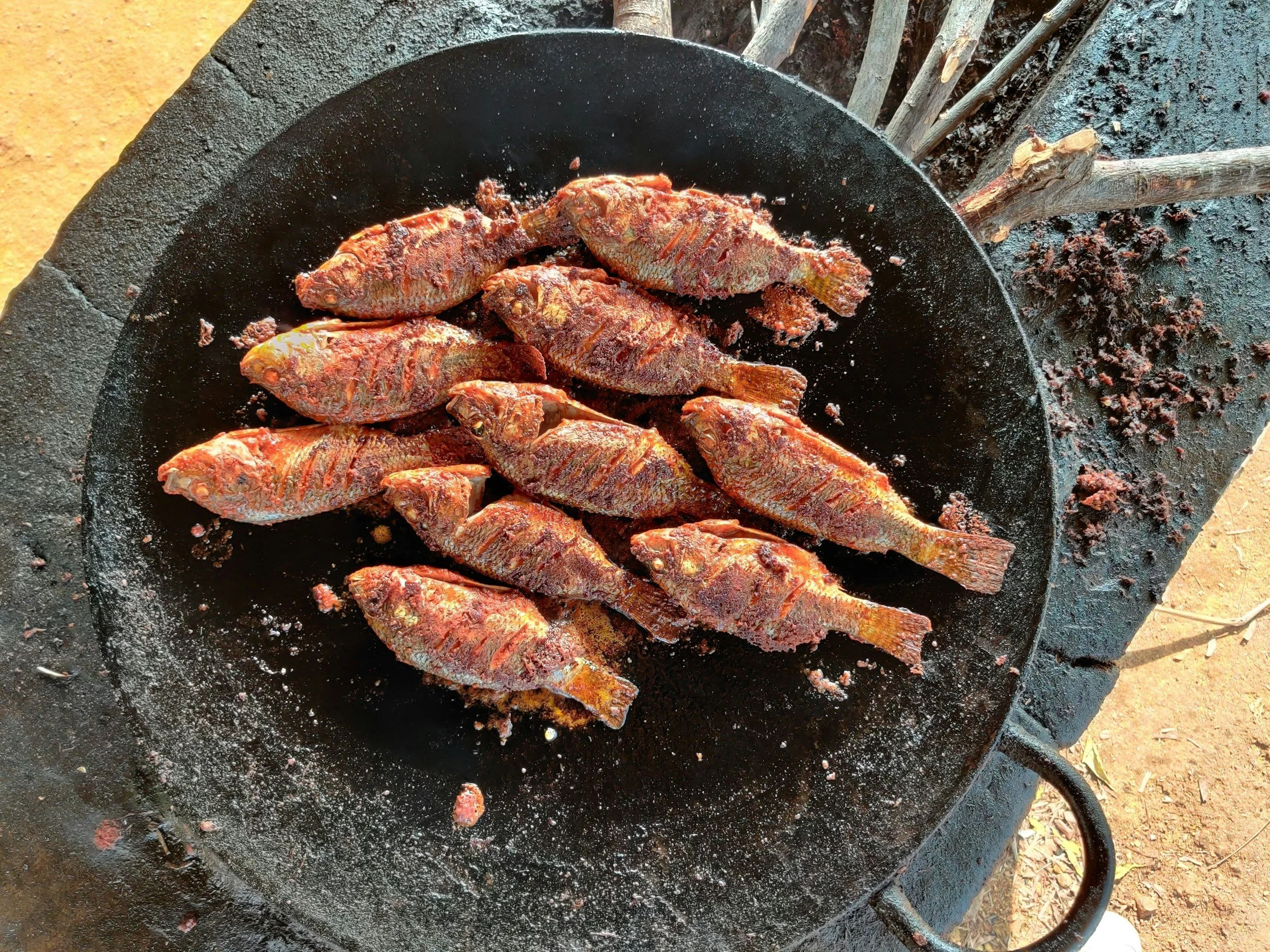 Fried fish being cooked on a black skillet over an open flame, with some herbs and sticks in the background.