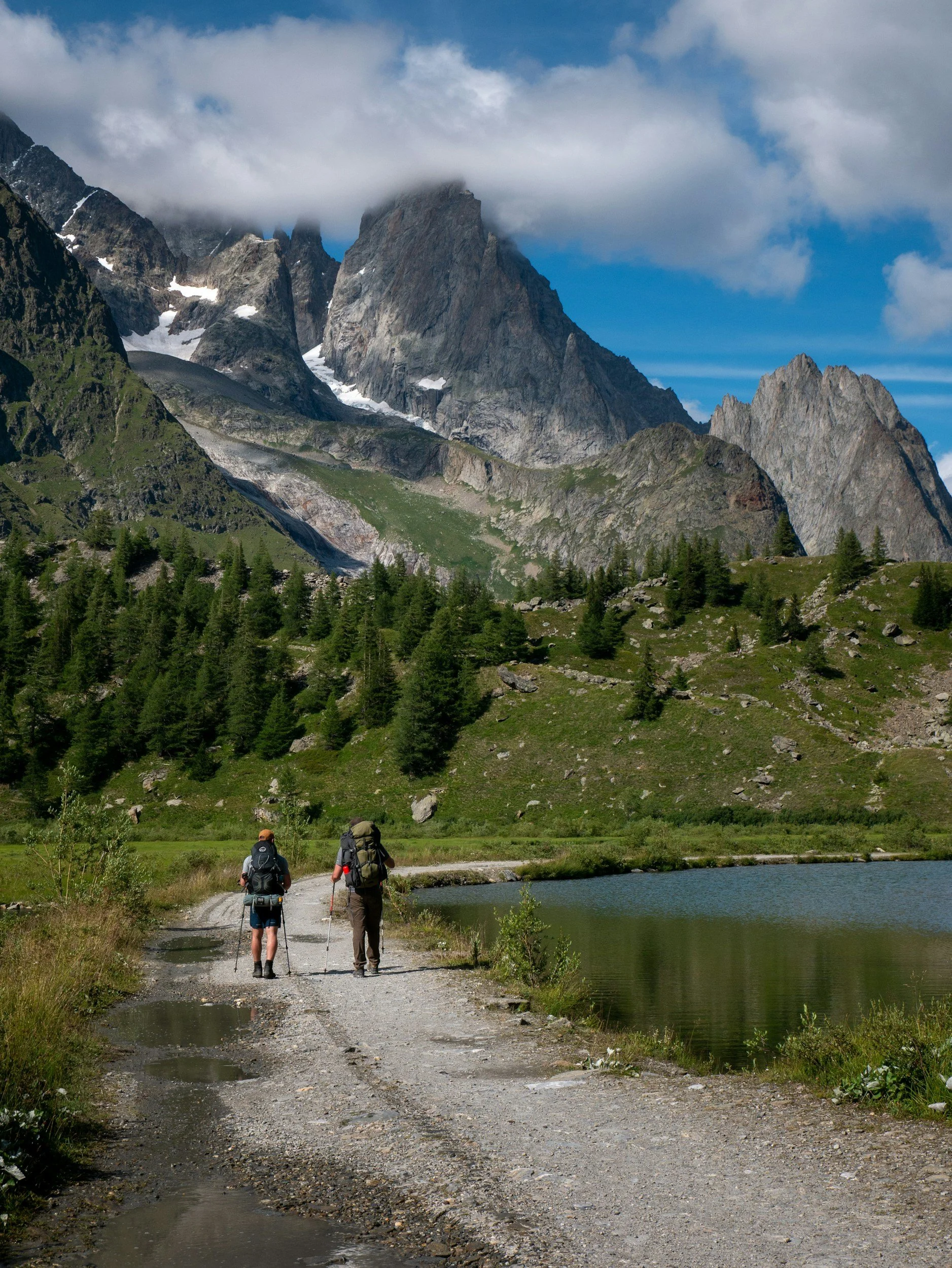 Two hikers walking along a gravel trail by a lake with green mountains and rocky peaks in the background under a partly cloudy sky.