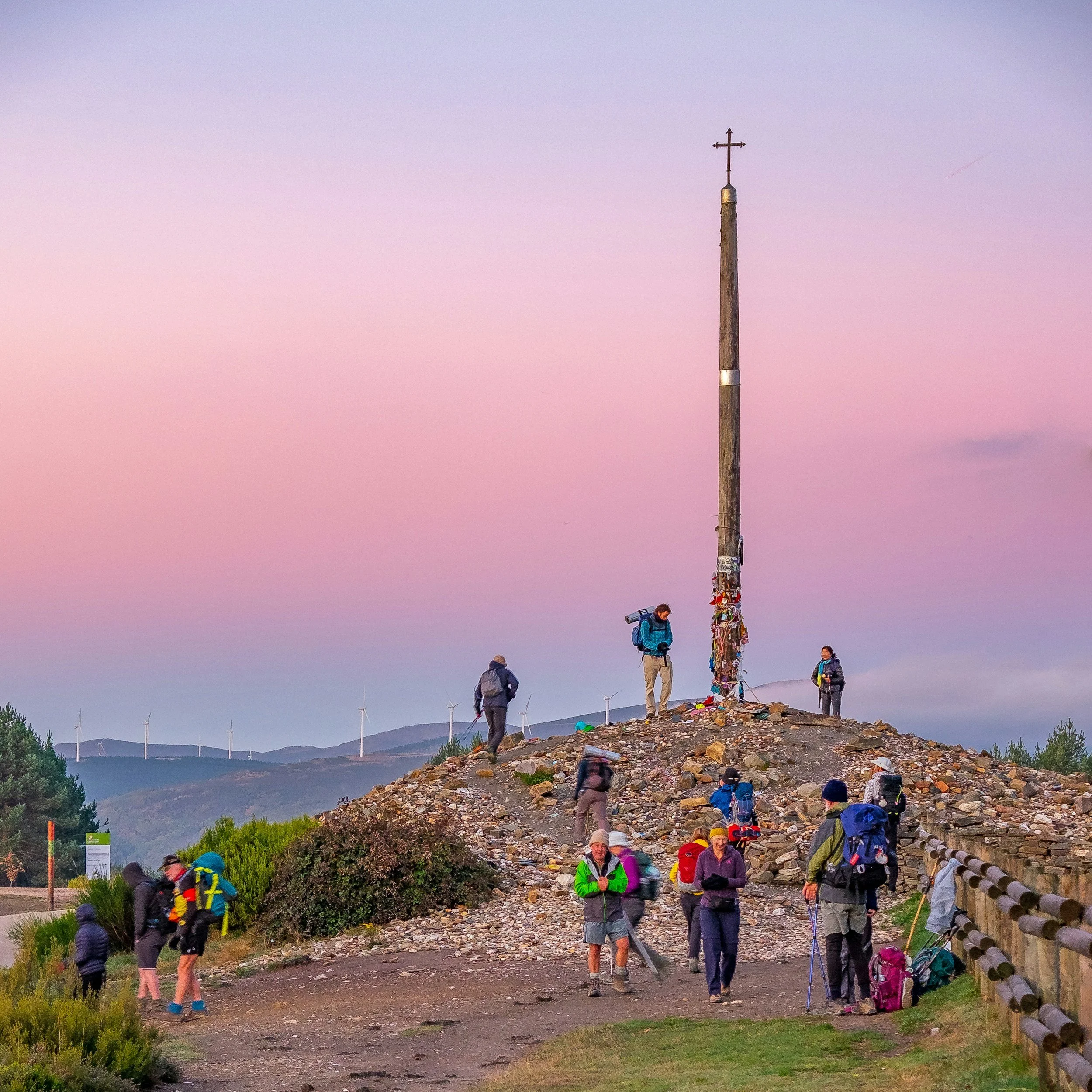 Group of hikers gathering around a tall wooden pole with a cross on top, on a rocky hill at sunset, with wind turbines visible in the distance.
