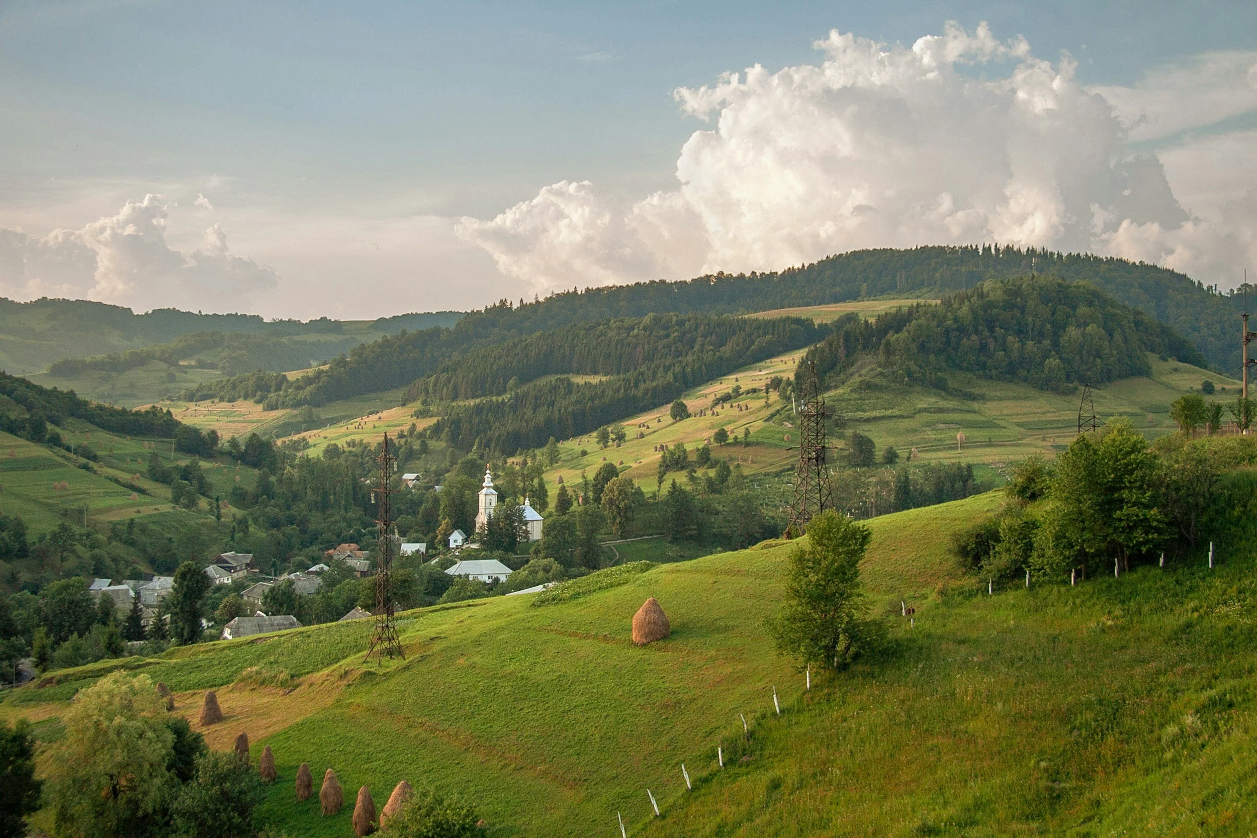 Green rolling hills with haystacks, trees, electricity pylons, and a village with a white church. Mountainous background under a partly cloudy sky.