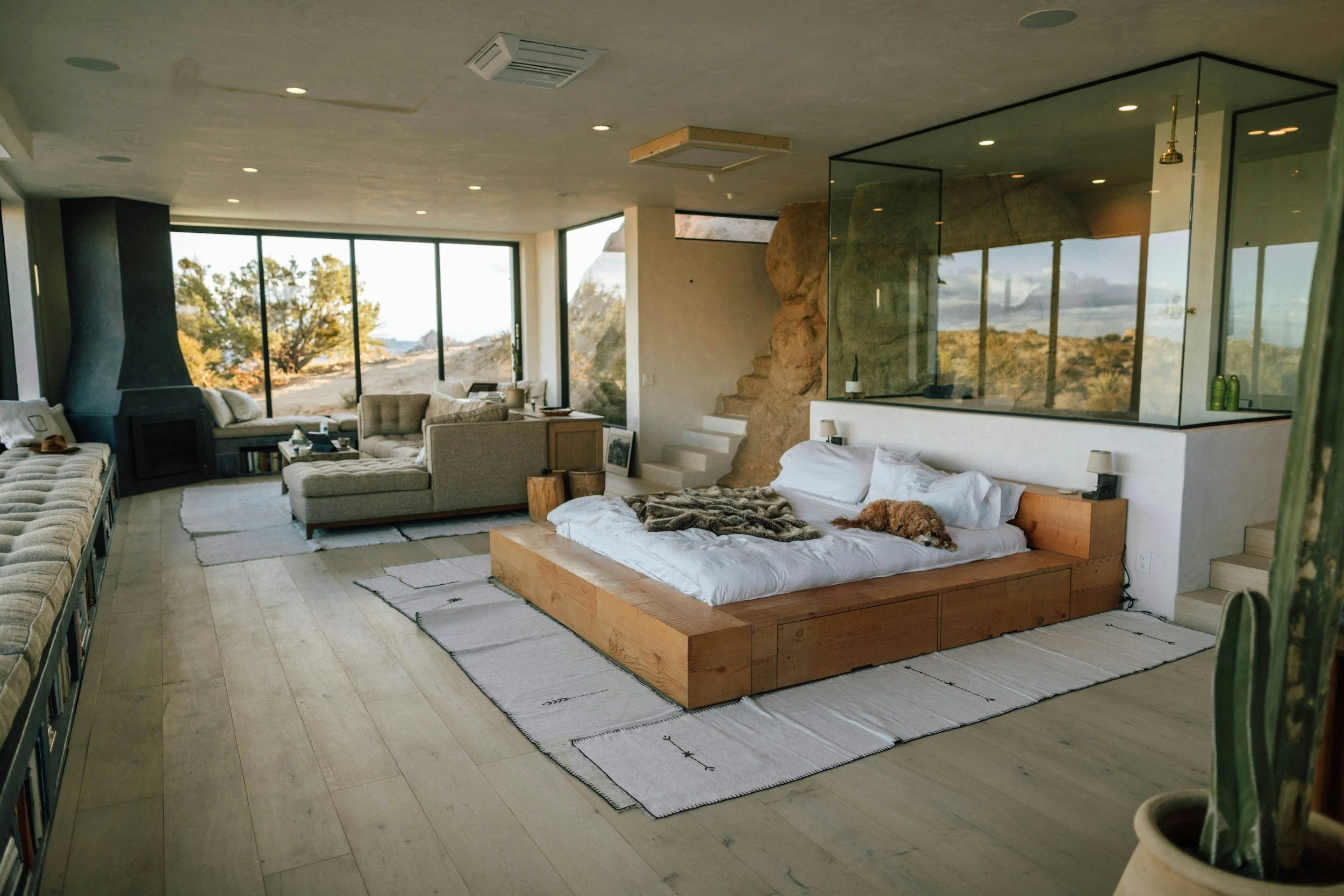 A modern bedroom with large windows showing a rocky landscape and trees outside. The room features a wooden bed with white linens and a small dog lying on it, and a seating area with beige couches, pillows, and a fireplace. There are rugs on the light wooden floor and a glass partition that separates the sleeping area from the rest of the room.