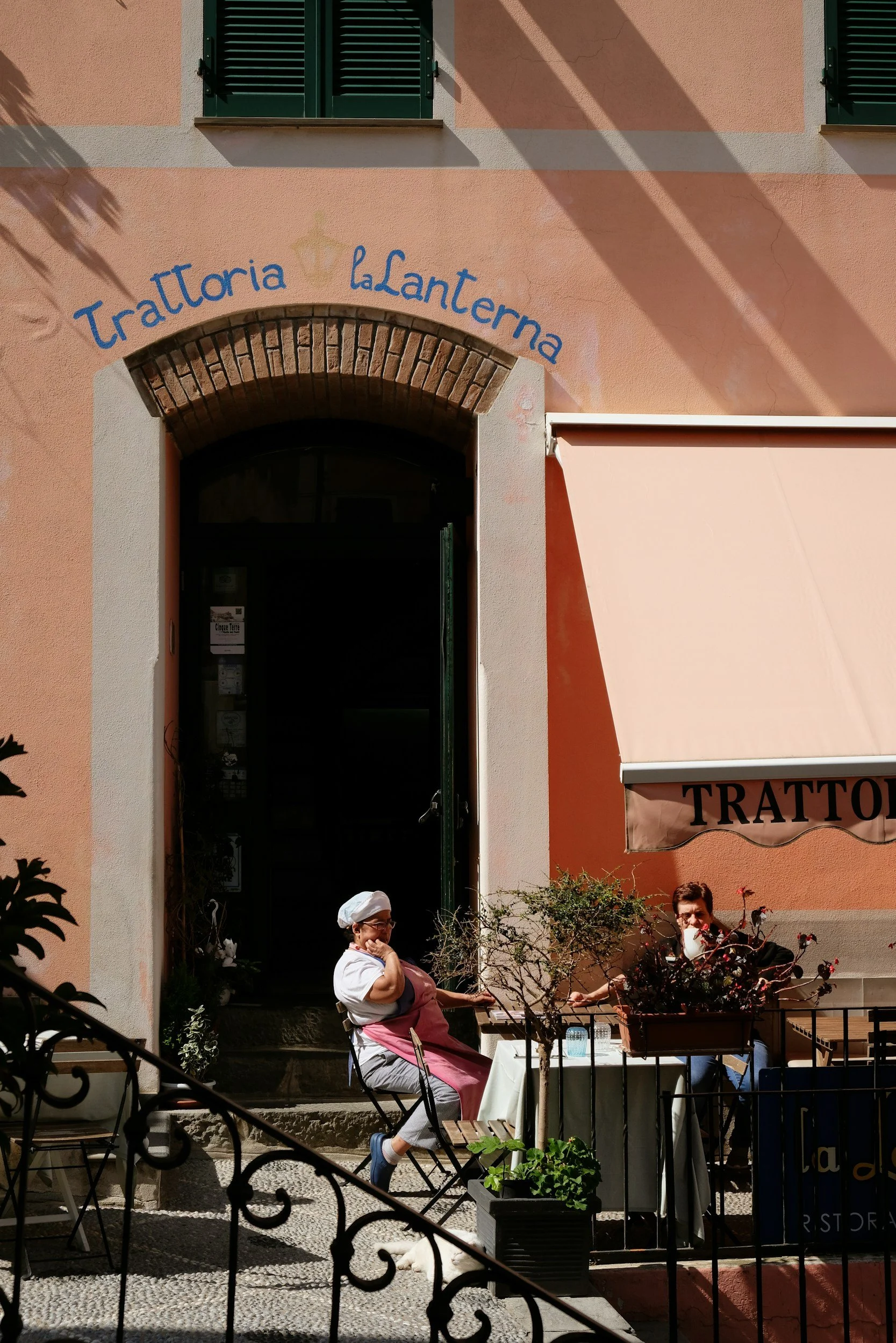 A woman sits outside a restaurant named Trattoria LaLanterna, wearing a pink apron and a chef's hat, with a second person sitting at the table. The restaurant has a pink exterior with green shutters and an awning. There are small trees and potted plants in the outdoor seating area.