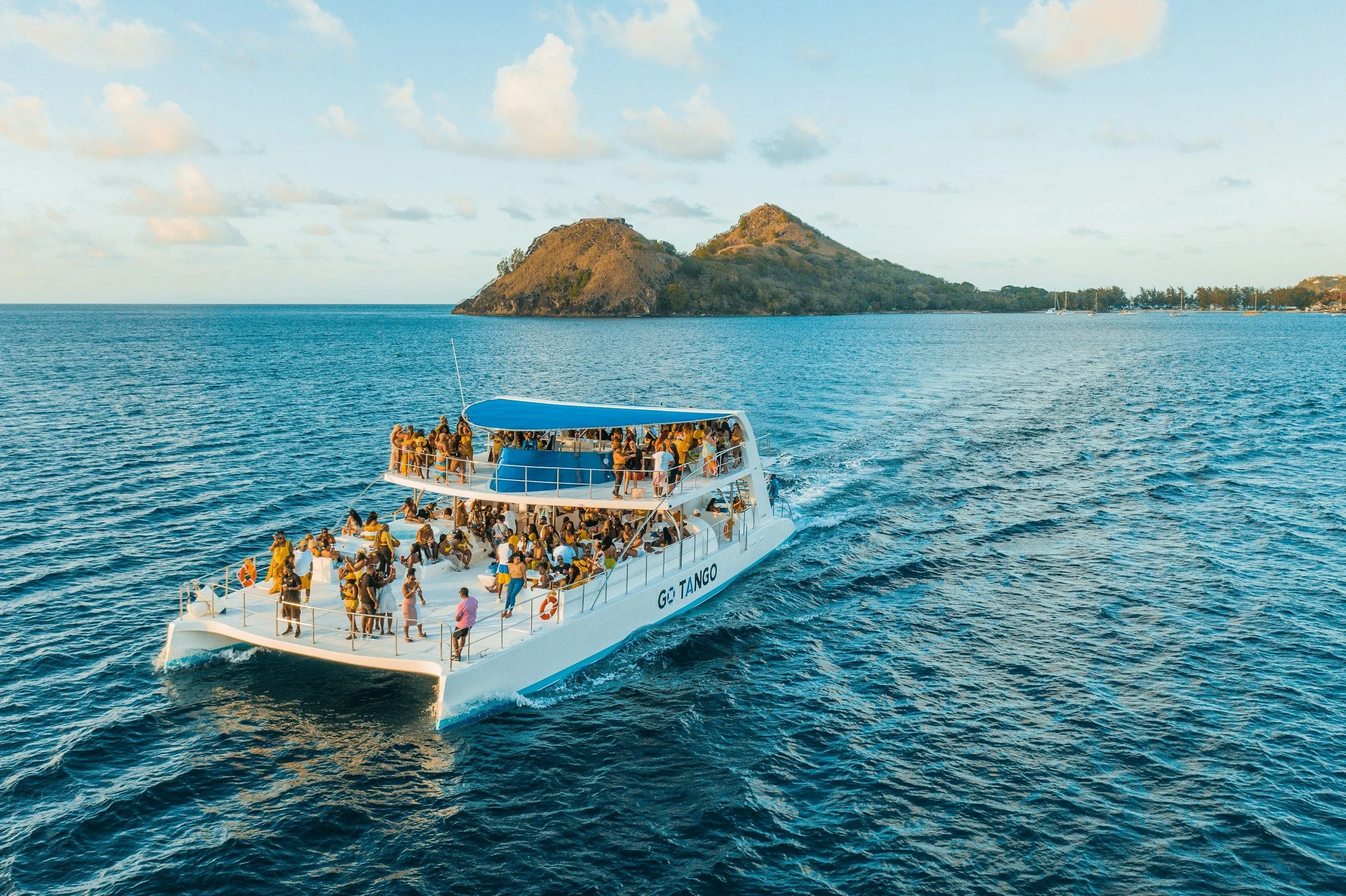 A large boat with many people onboard, cruising on blue water near an island with hills and trees, under partly cloudy sky.