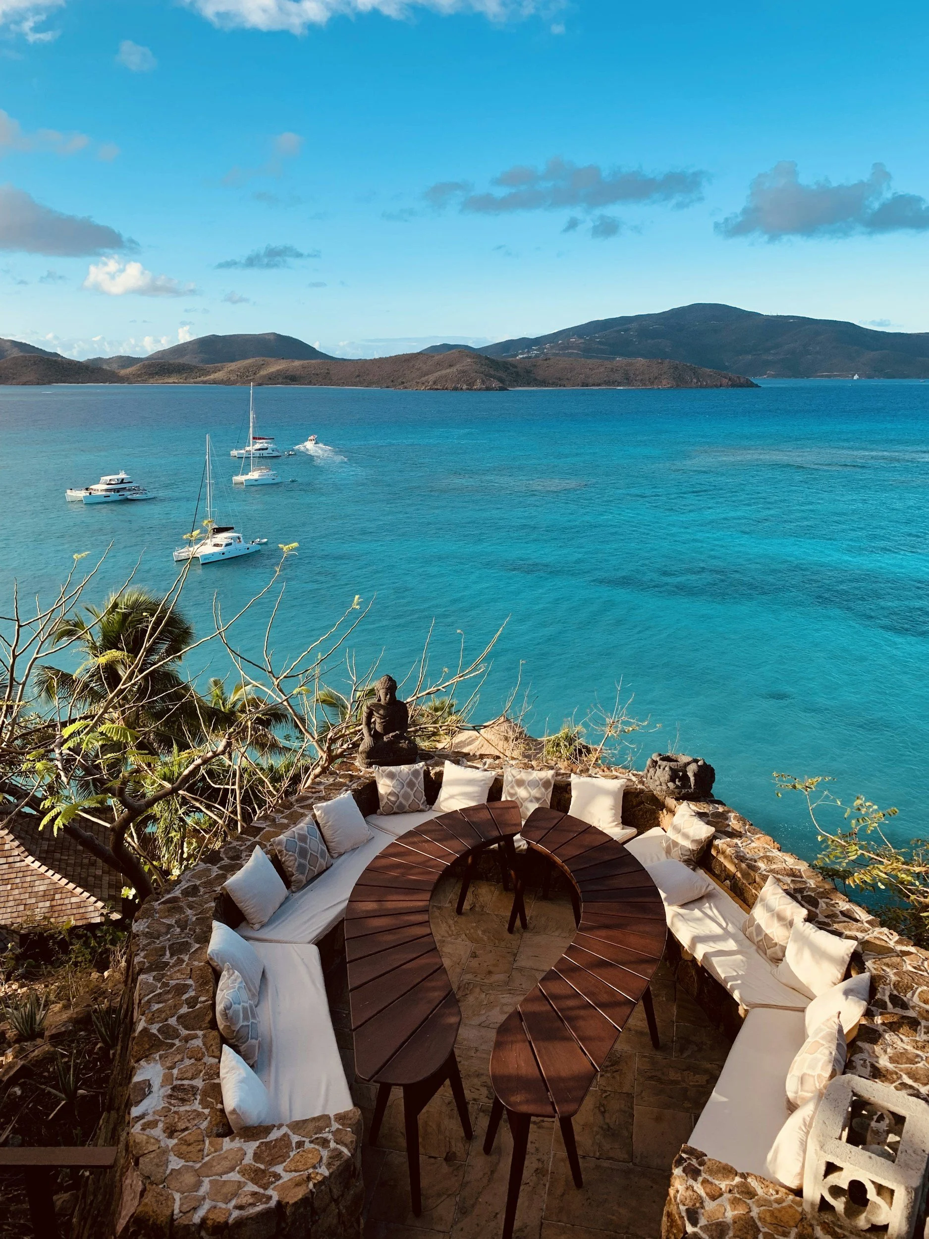 Outdoor seating area with a heart-shaped table, surrounded by cushioned benches and pillows, overlooking a tropical bay with sailboats and mountains in the background.
