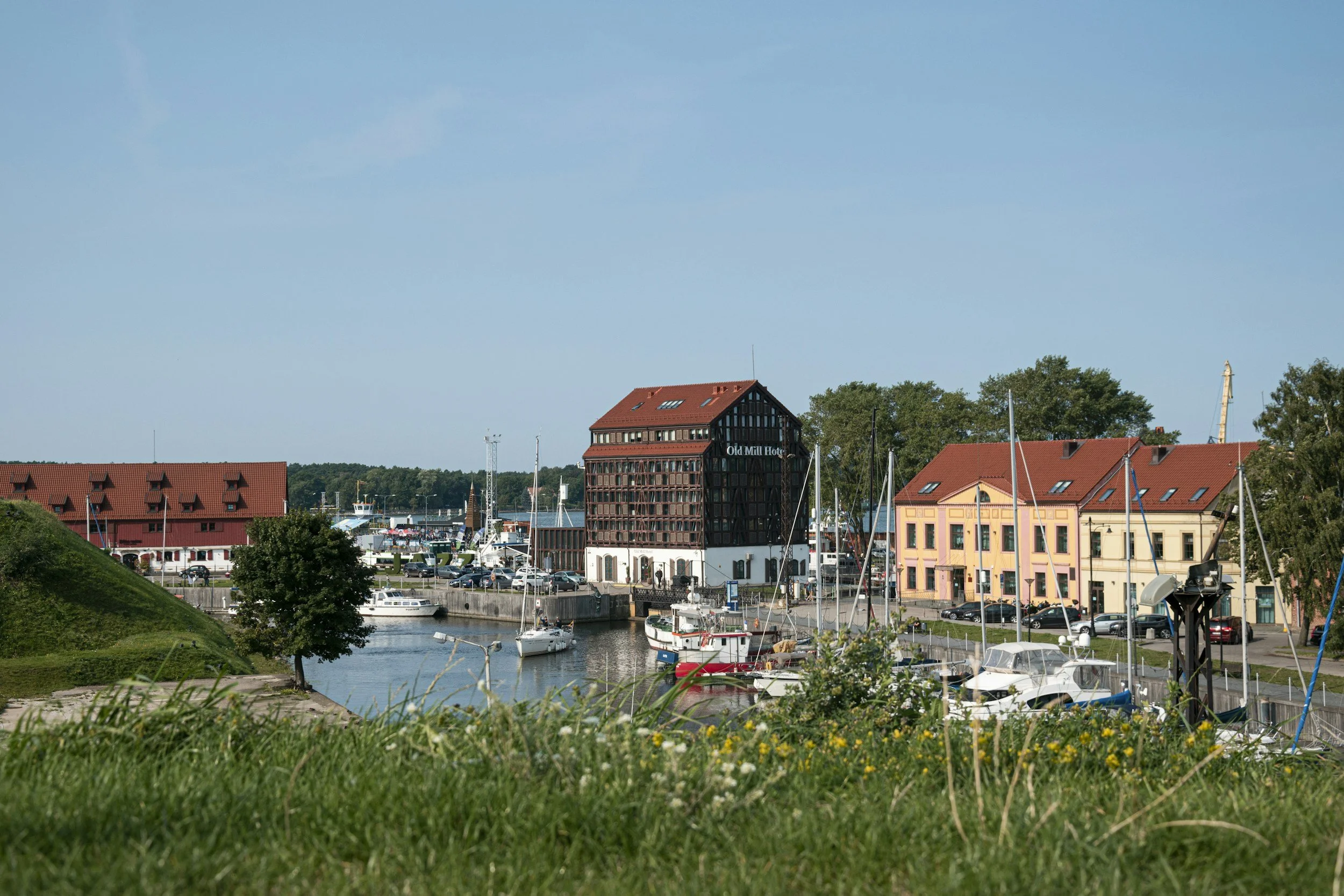 Harbor scene with boats docked and colorful buildings, including a dark wooden structure labeled 'Old Mill Hotel,' under a blue sky.