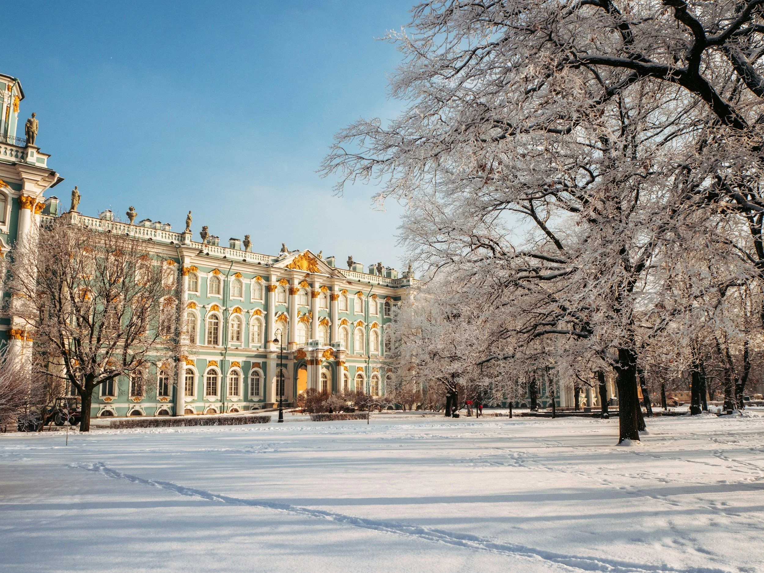 A historic building with ornate architecture, painted in green and gold, set against a winter landscape with snow-covered ground and trees, and footprints in the snow.