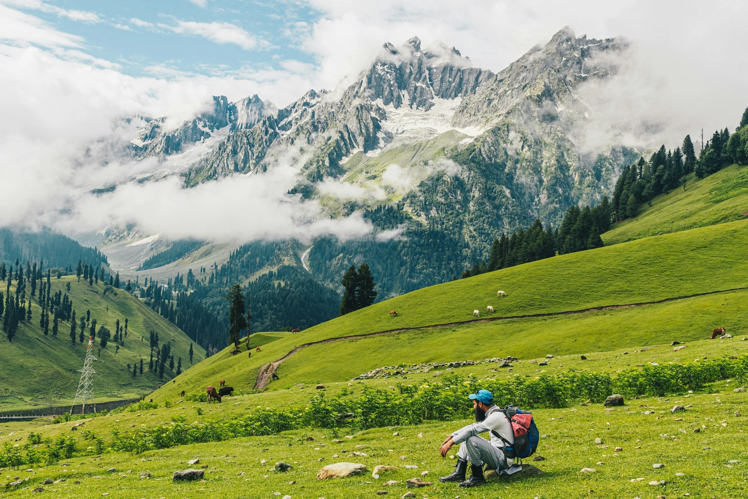 A man wearing outdoor gear and a backpack sitting on a grassy field, with green hills, trees, and snow-capped mountains in the background.