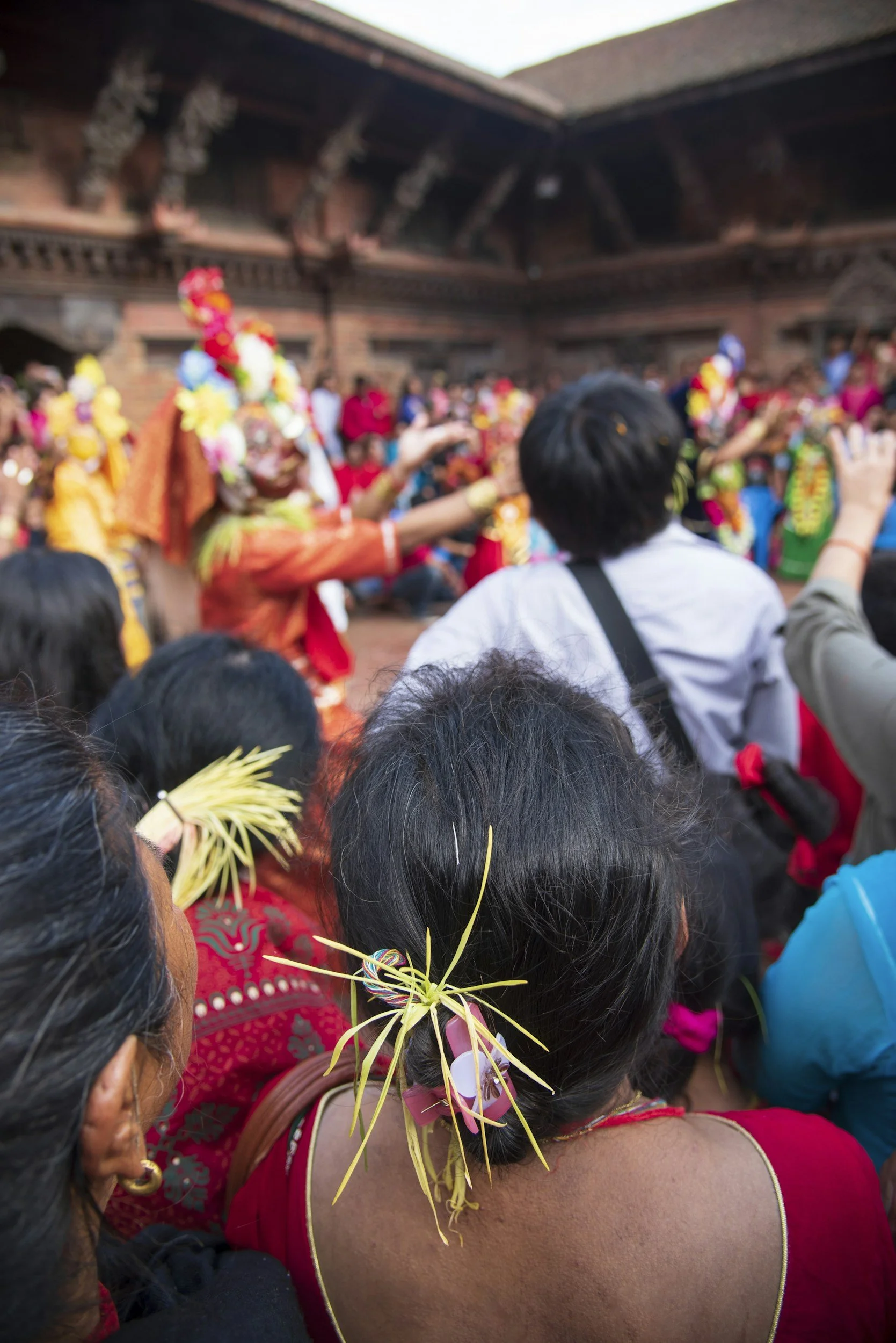People gathered at a cultural event, with some women dressed in traditional attire, wearing flower and palm decorations in their hair, participating in a dance or celebration in a courtyard.