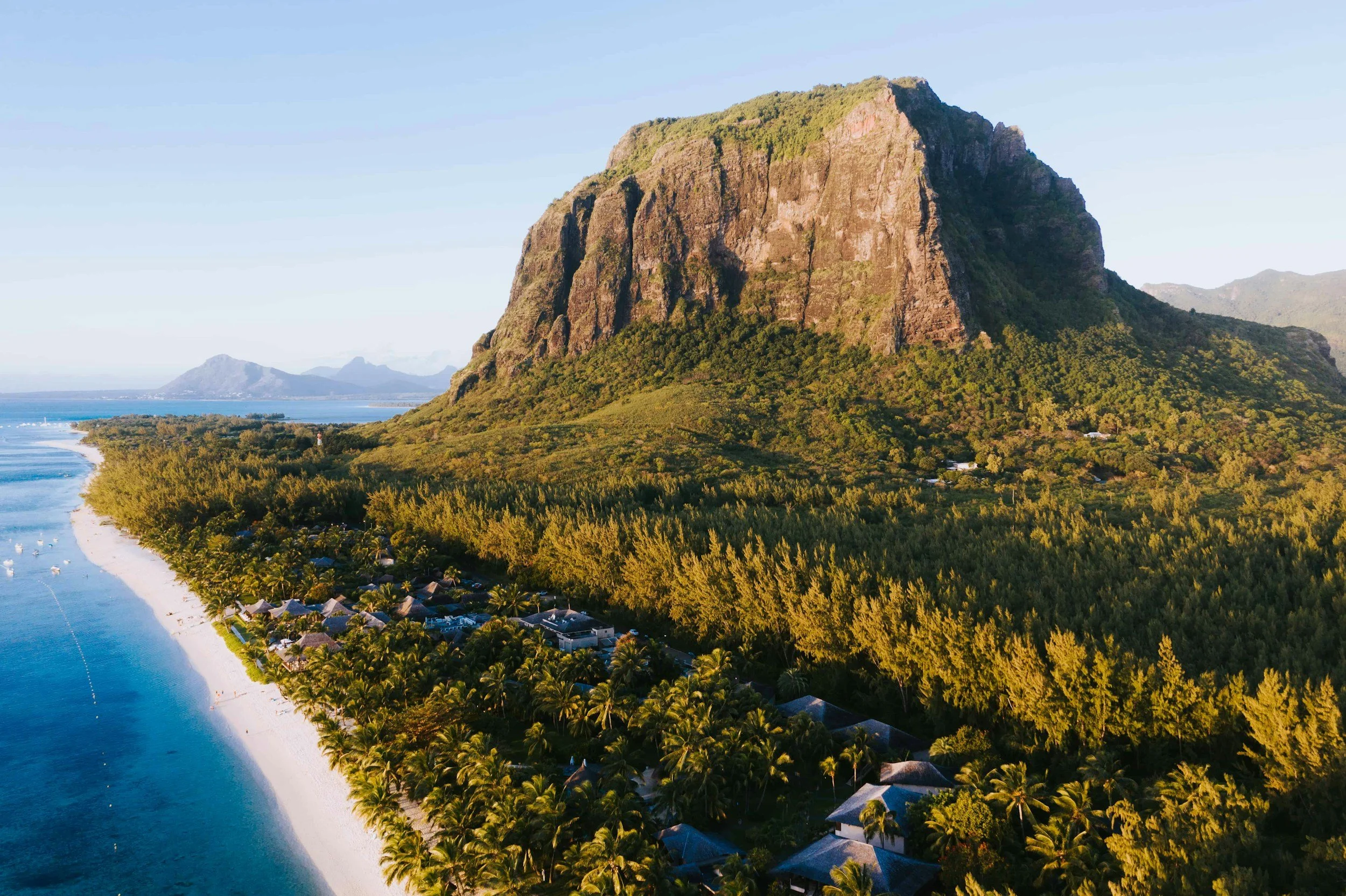 A coastal landscape with a large mountain, a beach lined with palm trees, and scattered buildings, during sunset.
