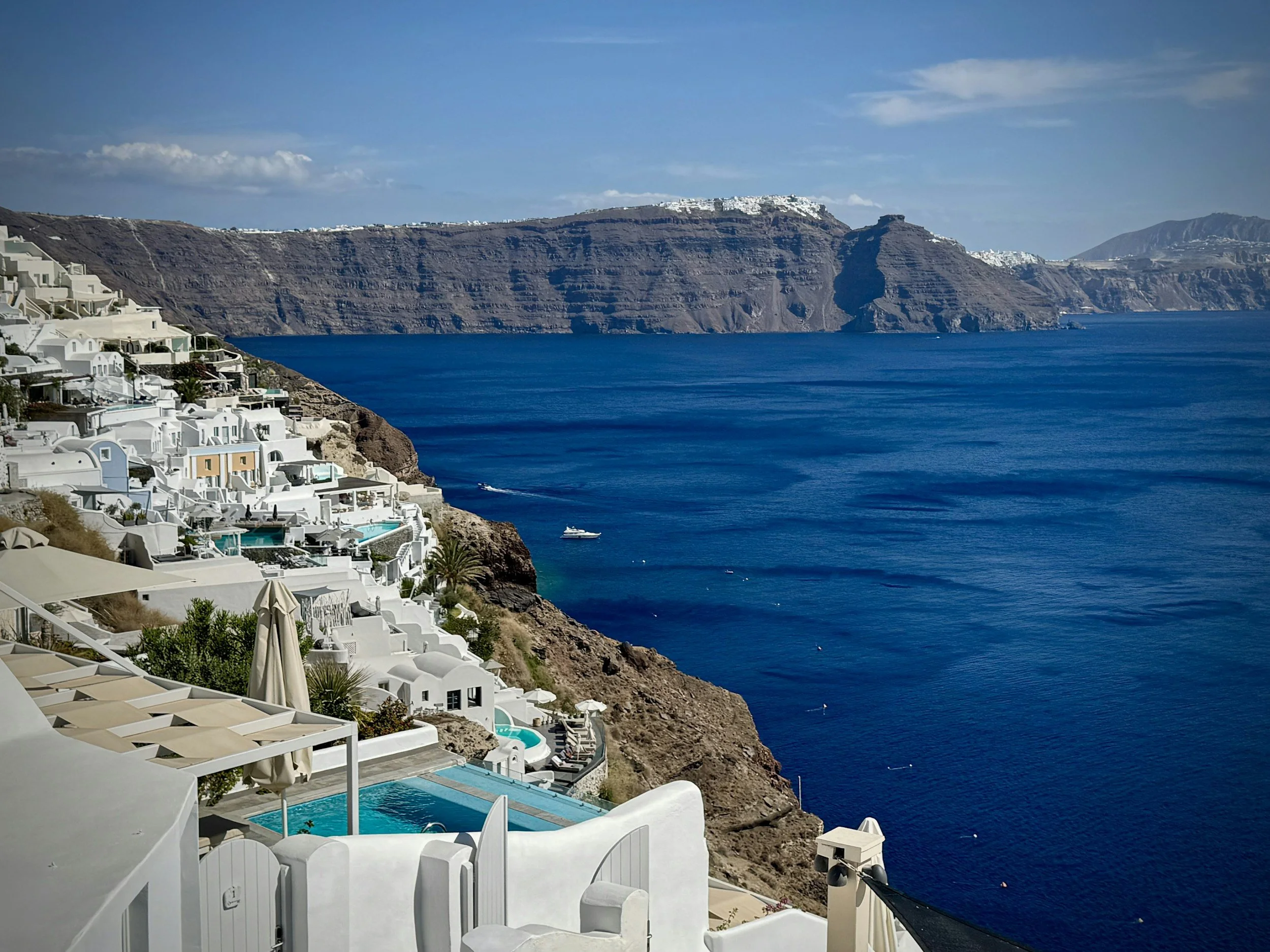 White buildings with terraces and pools on a cliffside overlooking the deep blue sea, with a mountainous island in the distance.