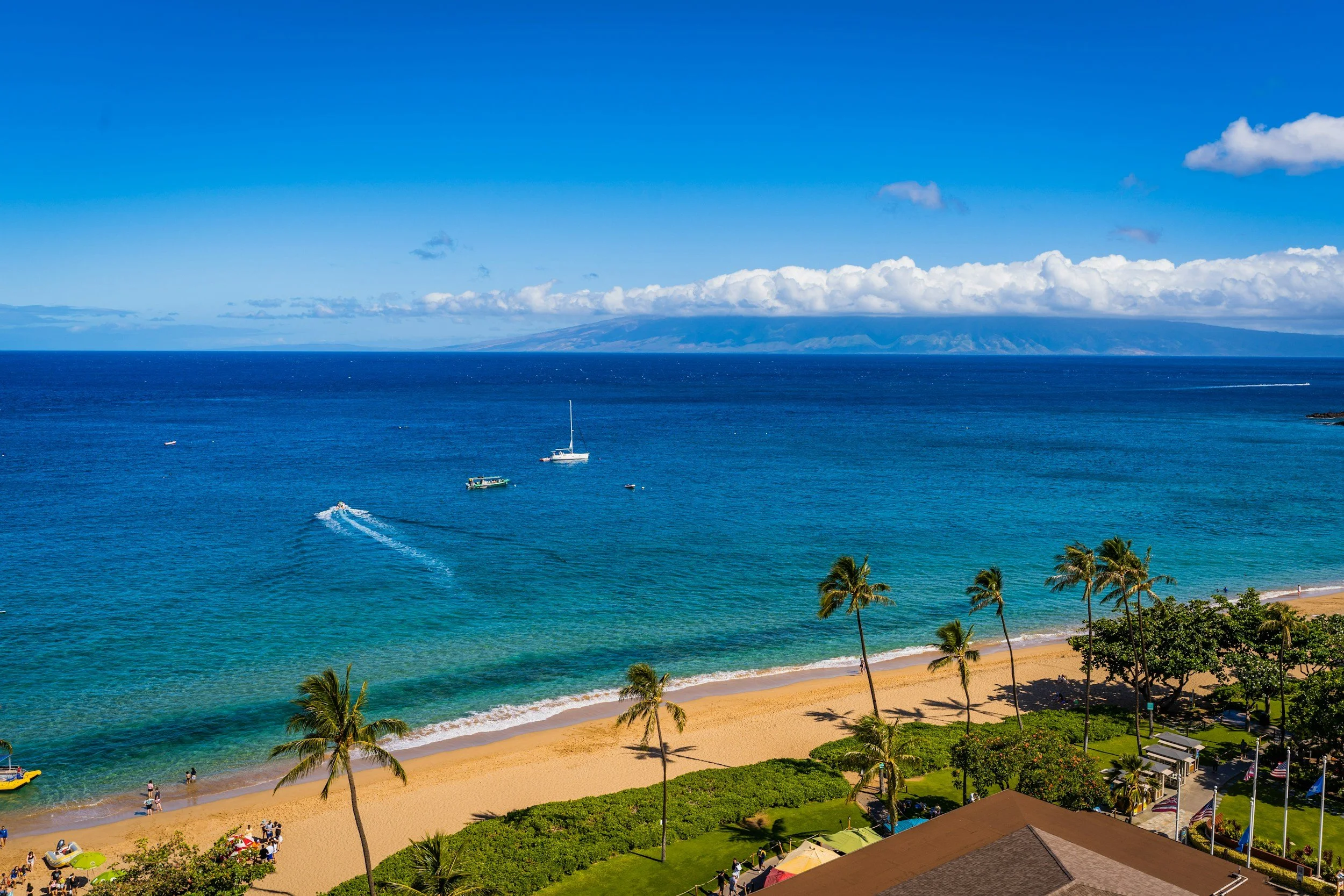 A tropical beach scene with golden sand, palm trees, and green foliage in the foreground. Several people are near the water, some standing on the shore. The ocean is a vibrant blue with a few boats and a sailboat, and a mountain range is visible in the distance under a partly cloudy sky.