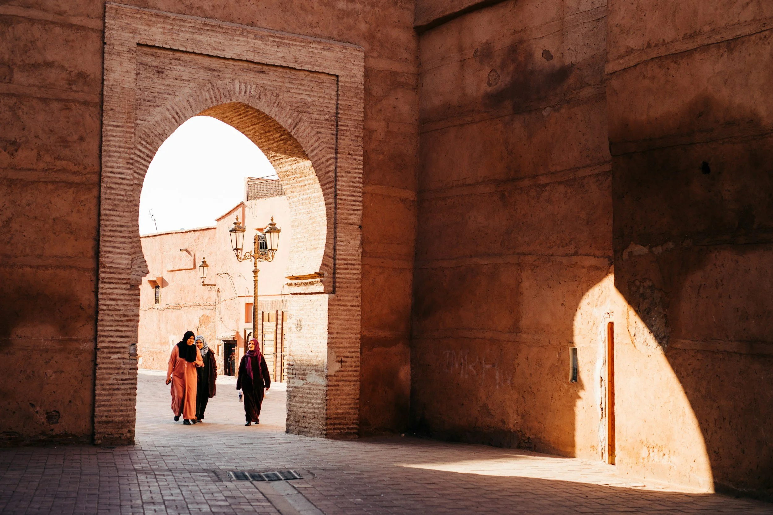 Three women walking through a historic gate in a desert city, with pinkish walls, a streetlamp, and bright sunlight casting shadows.
