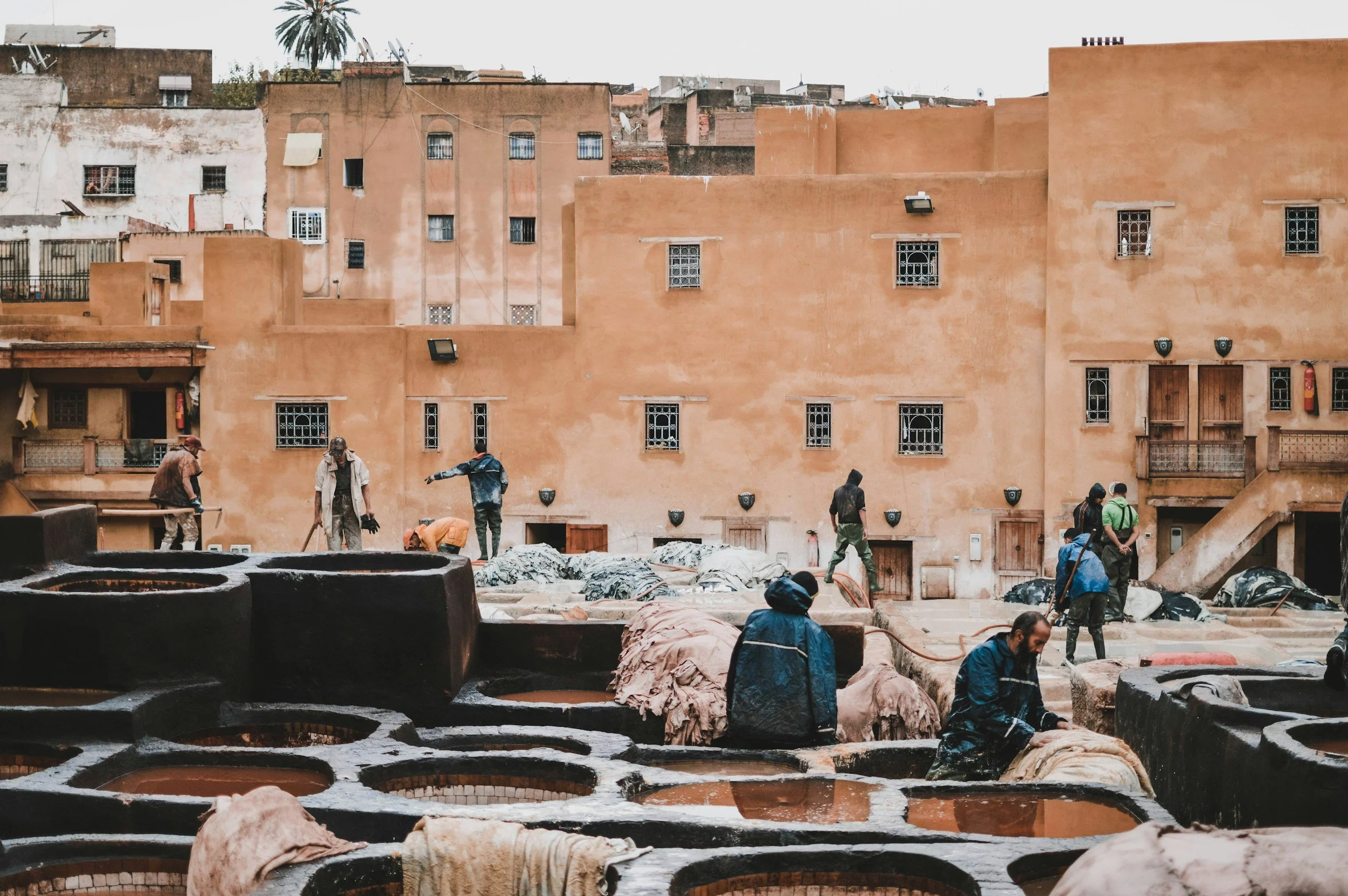 People working at a traditional tannery with large stone containers filled with liquids, surrounded by old buildings with small windows in the background.