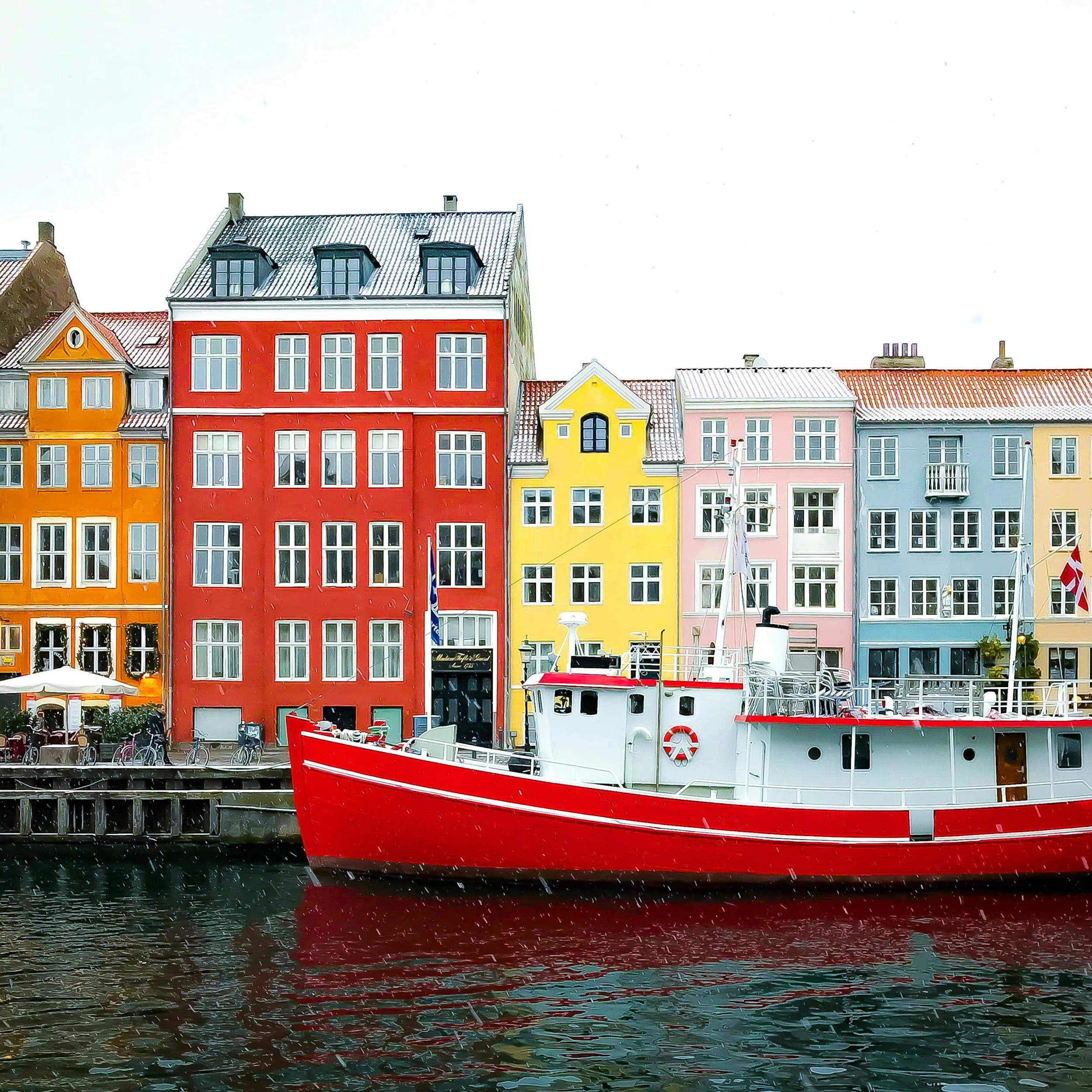 Colorful waterfront buildings with a red boat docked in front, in a city scene with bright colors and a cloudy sky.