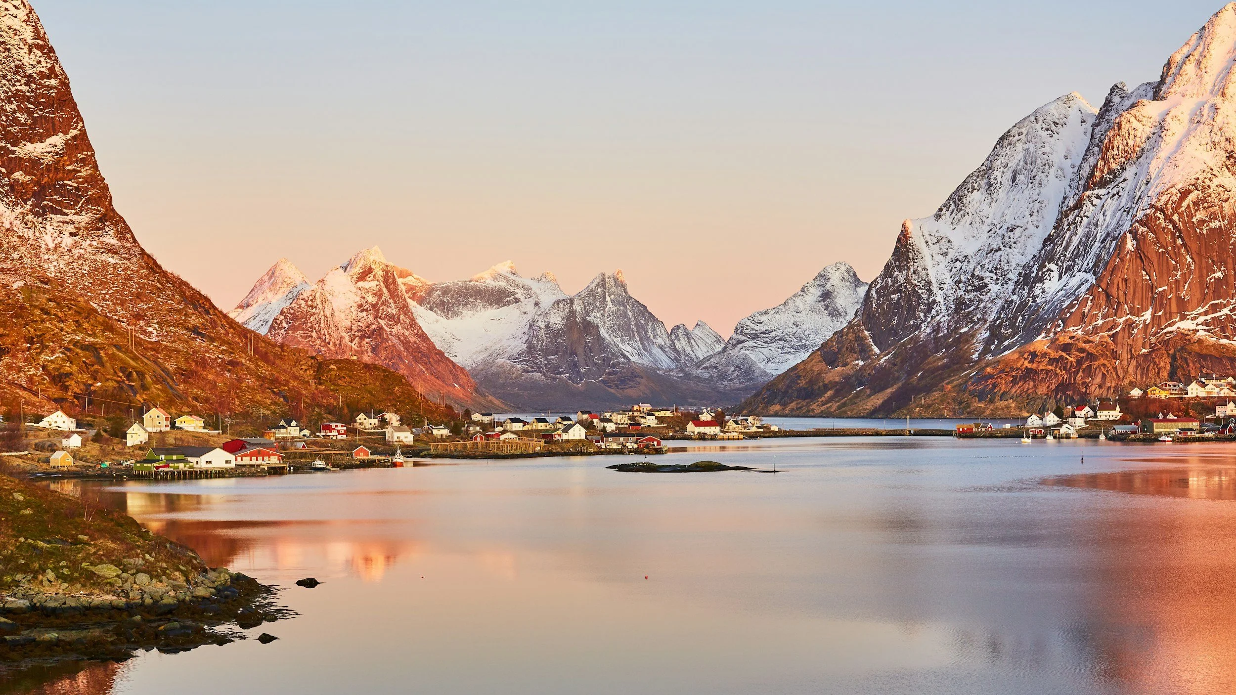 Scenic view of snow-capped mountains surrounding a calm water body with a small village of colorful houses along the shore at sunset.