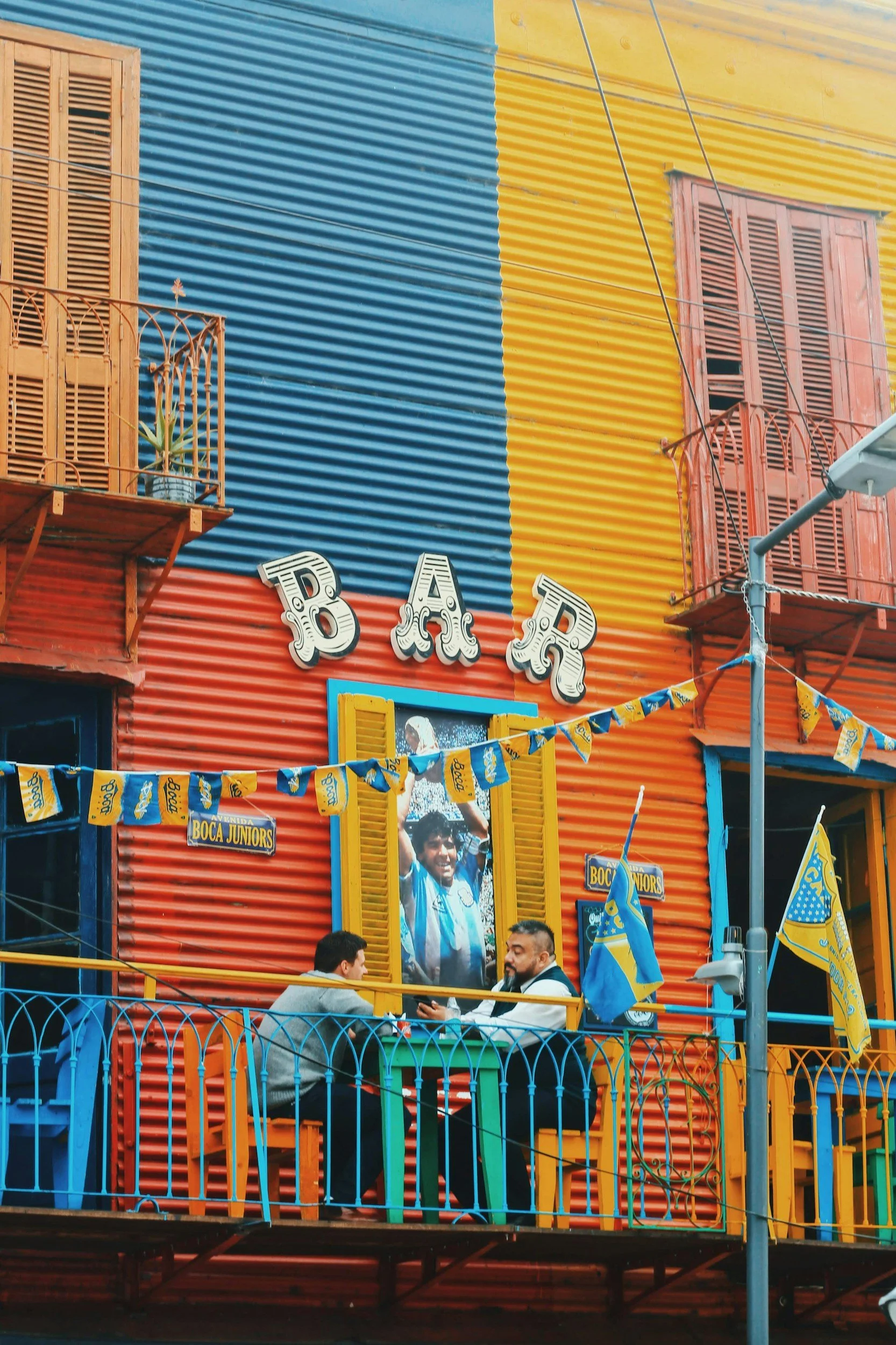 Colorful building with yellow, red, blue, and orange corrugated metal walls, featuring open wooden shutters and small balconies. Two men are sitting at a table on an outdoor balcony, with a large framed photo of a smiling man in a blue and white sports jersey hanging on the wall behind them. The scene is decorated with flags and banners, including ones with the Boca Juniors soccer team logo.