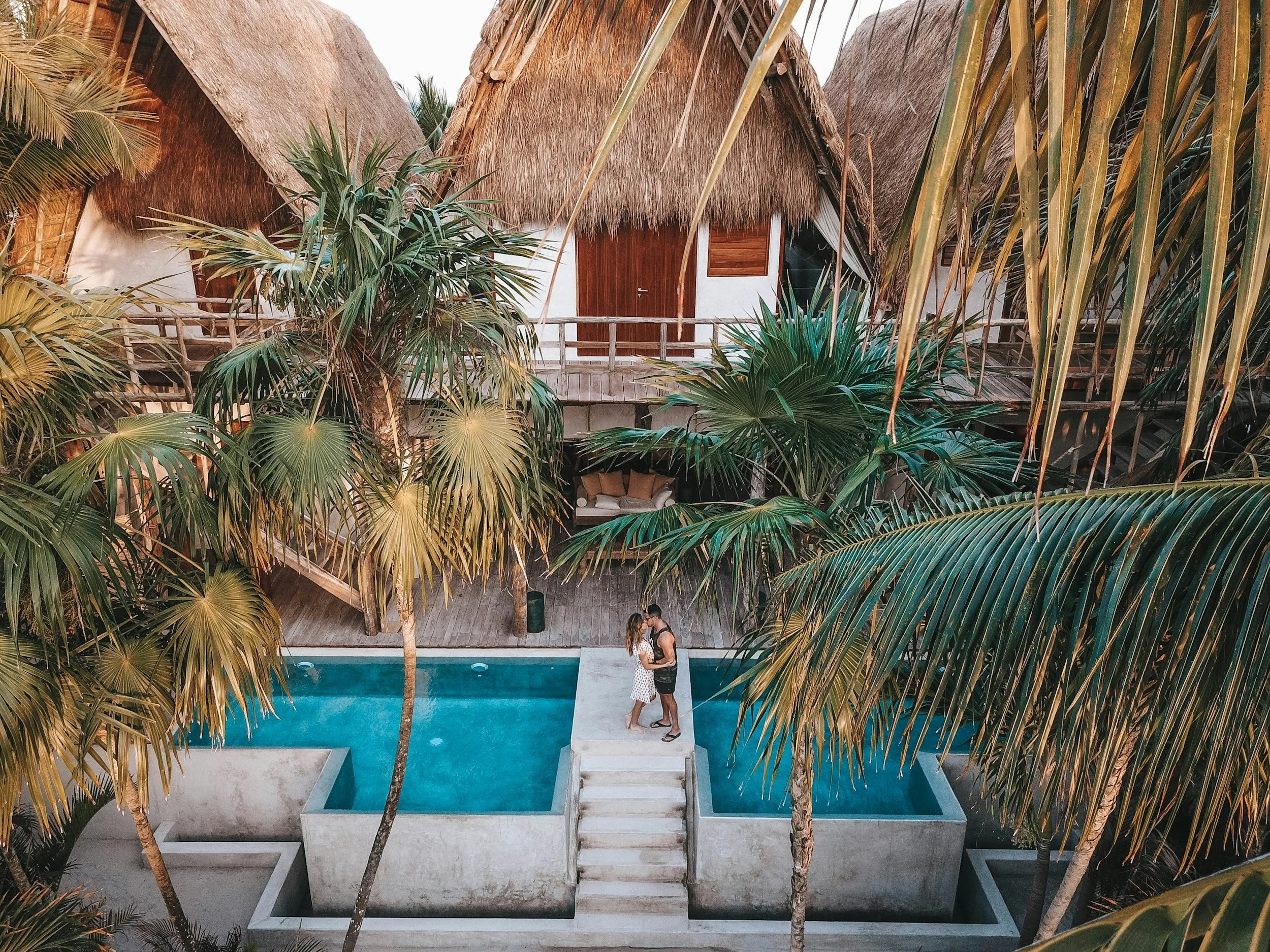 Couple standing on a small bridge over a pool at a tropical resort with palm trees and thatched roofs.