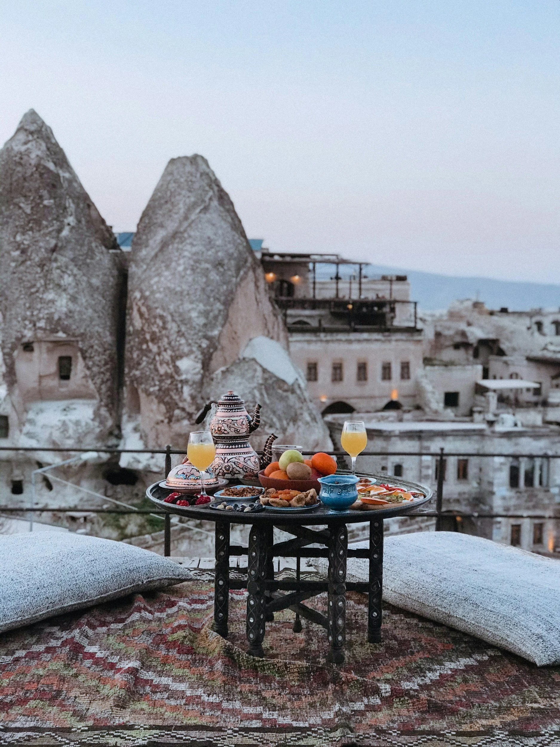 A rooftop breakfast setup with a small table, pillows, and cushions overlooking rock formations and buildings in Cappadocia, Turkey, featuring a teapot, two glasses of orange juice, and various breakfast foods.