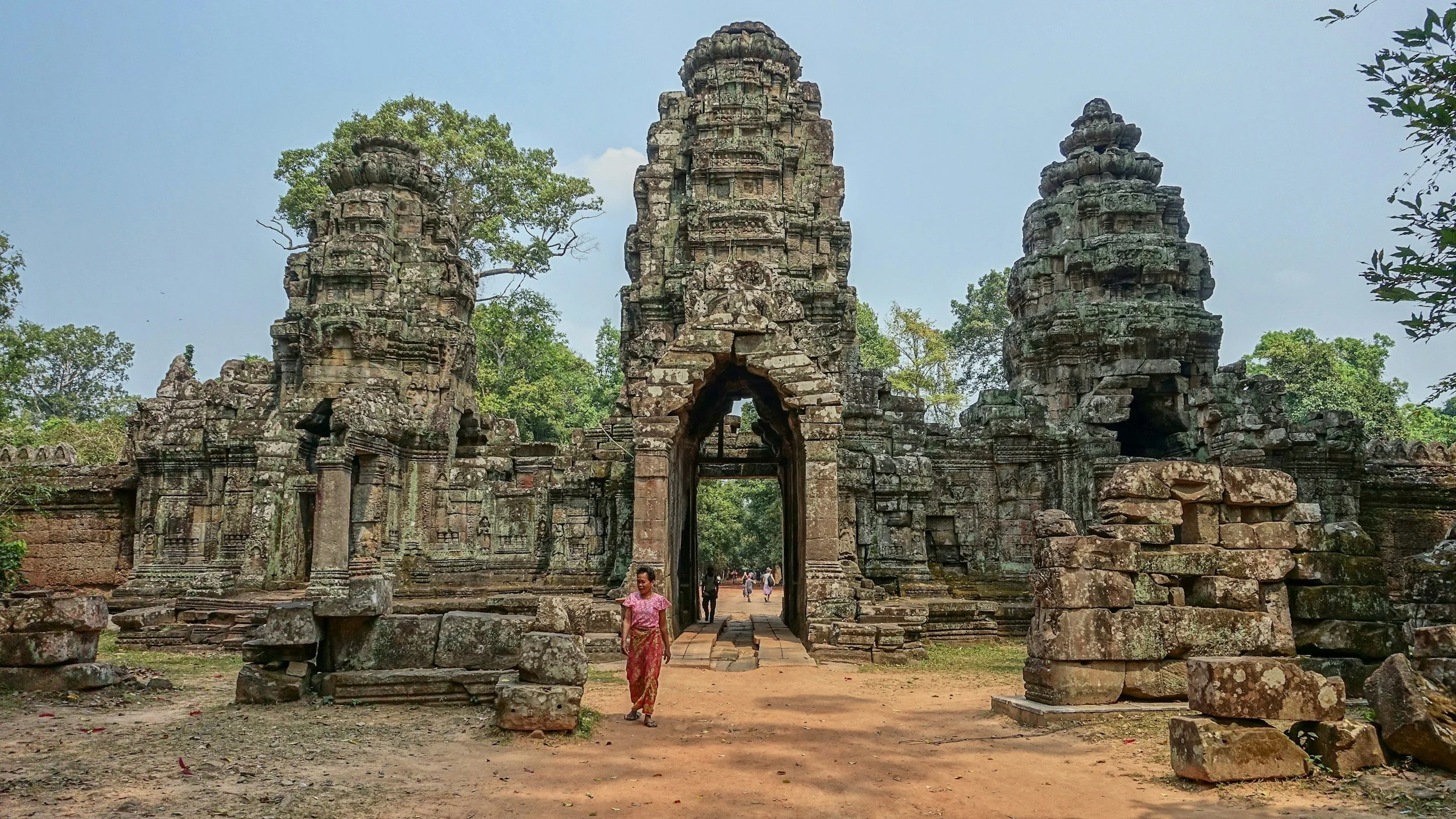 Ancient stone temple gate with three tall towers, surrounded by trees and ruins, a person walking through the entrance on a dirt pathway.