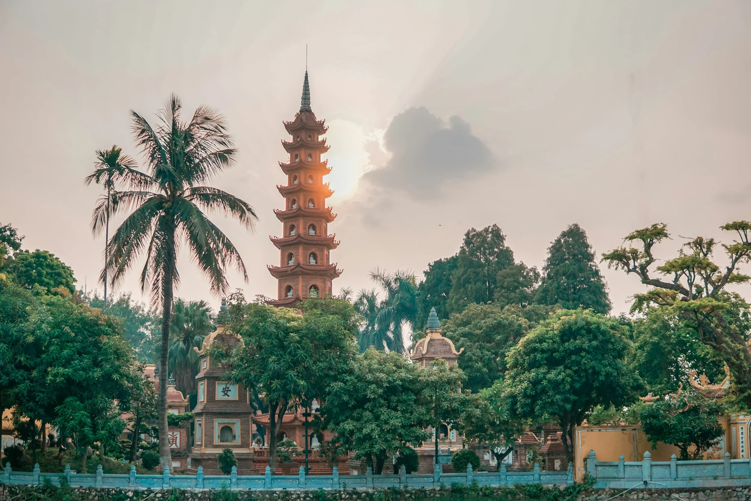 A traditional Asian pagoda surrounded by trees and greenery, with the sun partially visible behind it, in a lush garden setting.