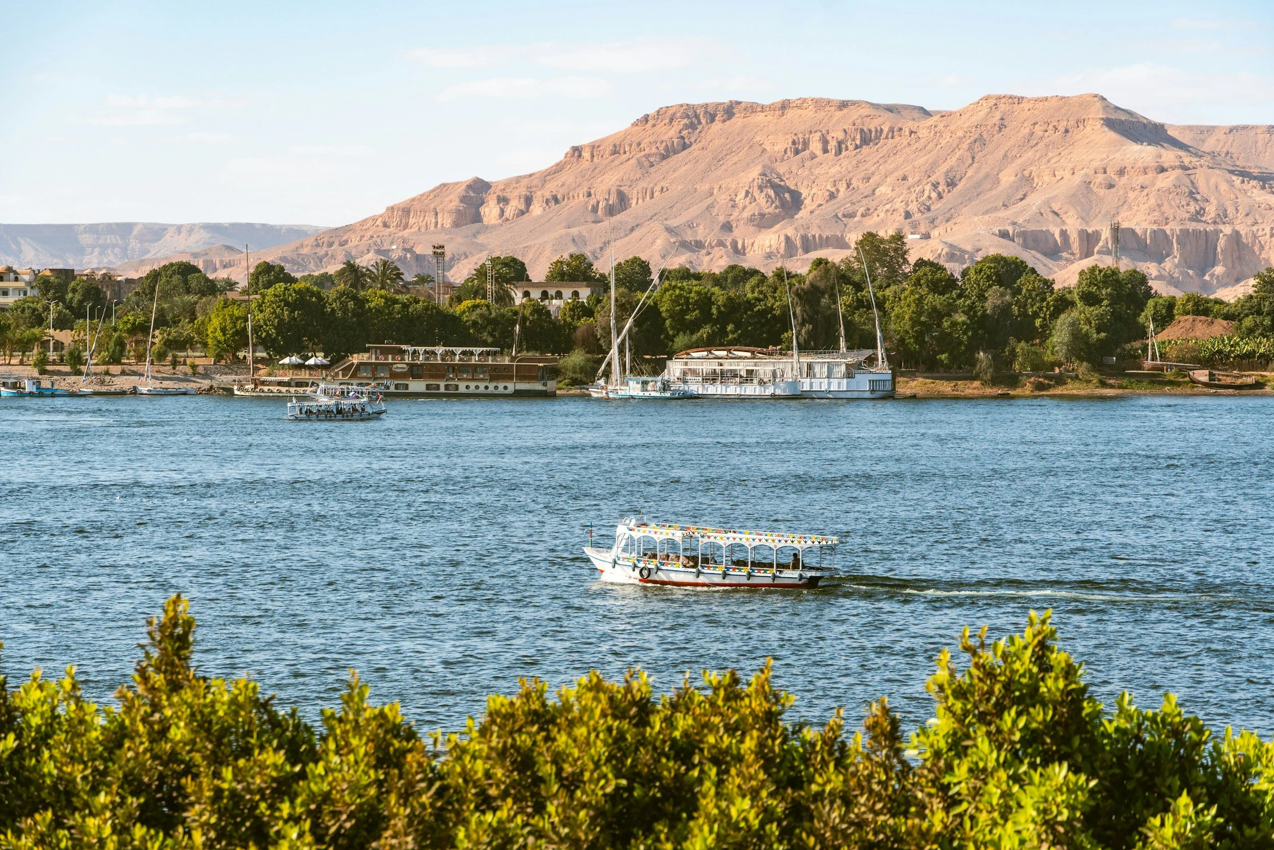 View of a large white cruise ship and some sailboats on a river, with ancient Egyptian-style buildings and temples in the background under a clear blue sky.