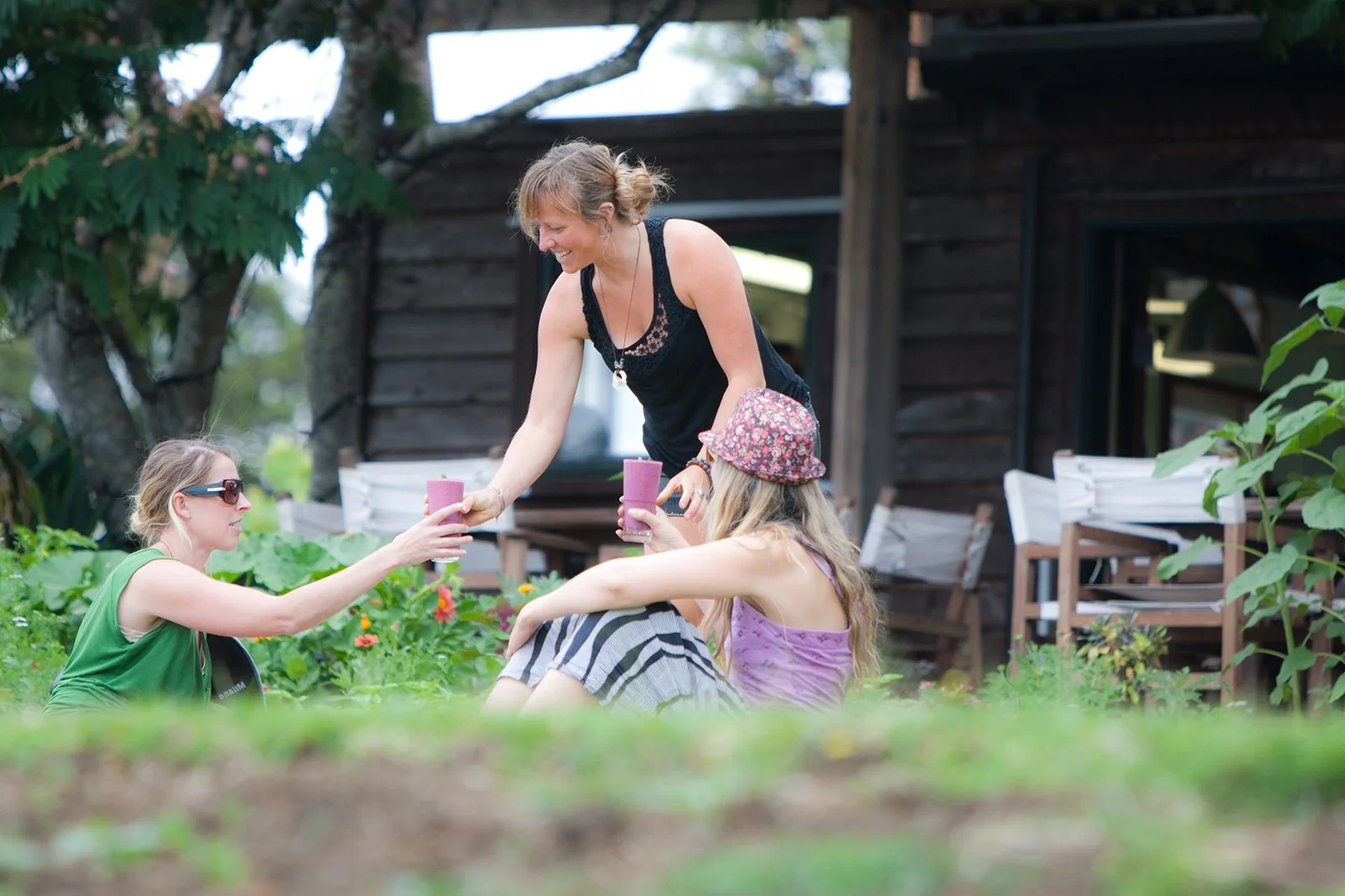 Three women outdoors, two sitting on the grass and one standing, exchanging drinks, with garden and wooden house in background.