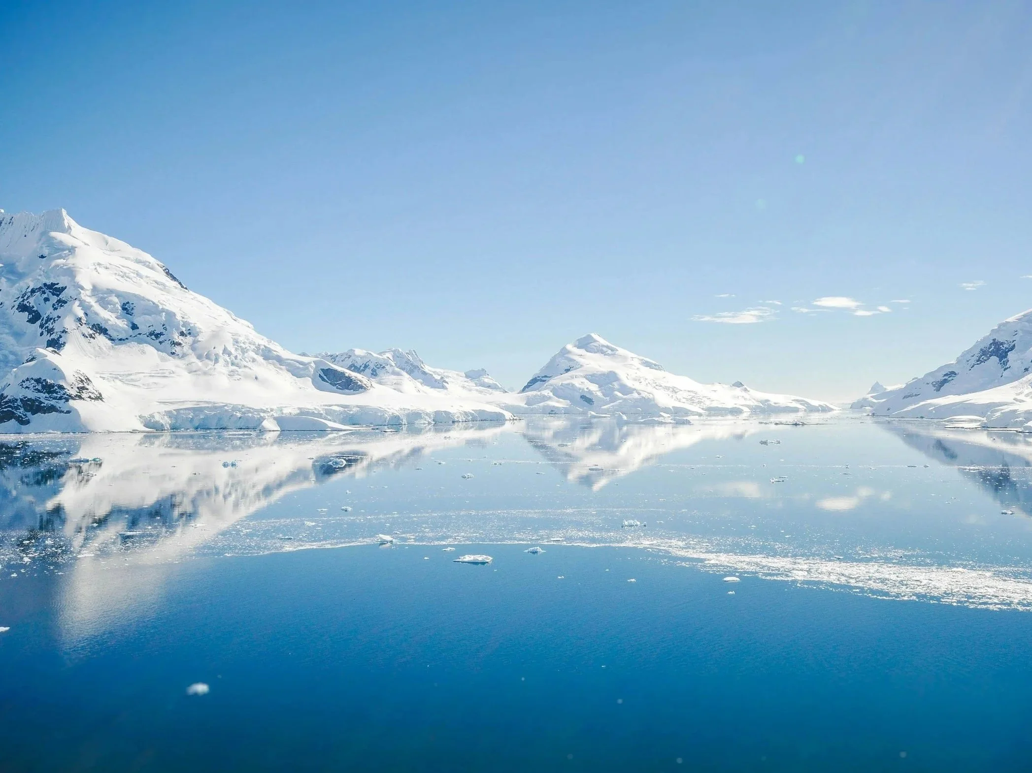 Bright blue sky over snow-covered mountains and calm reflective water with floating ice.