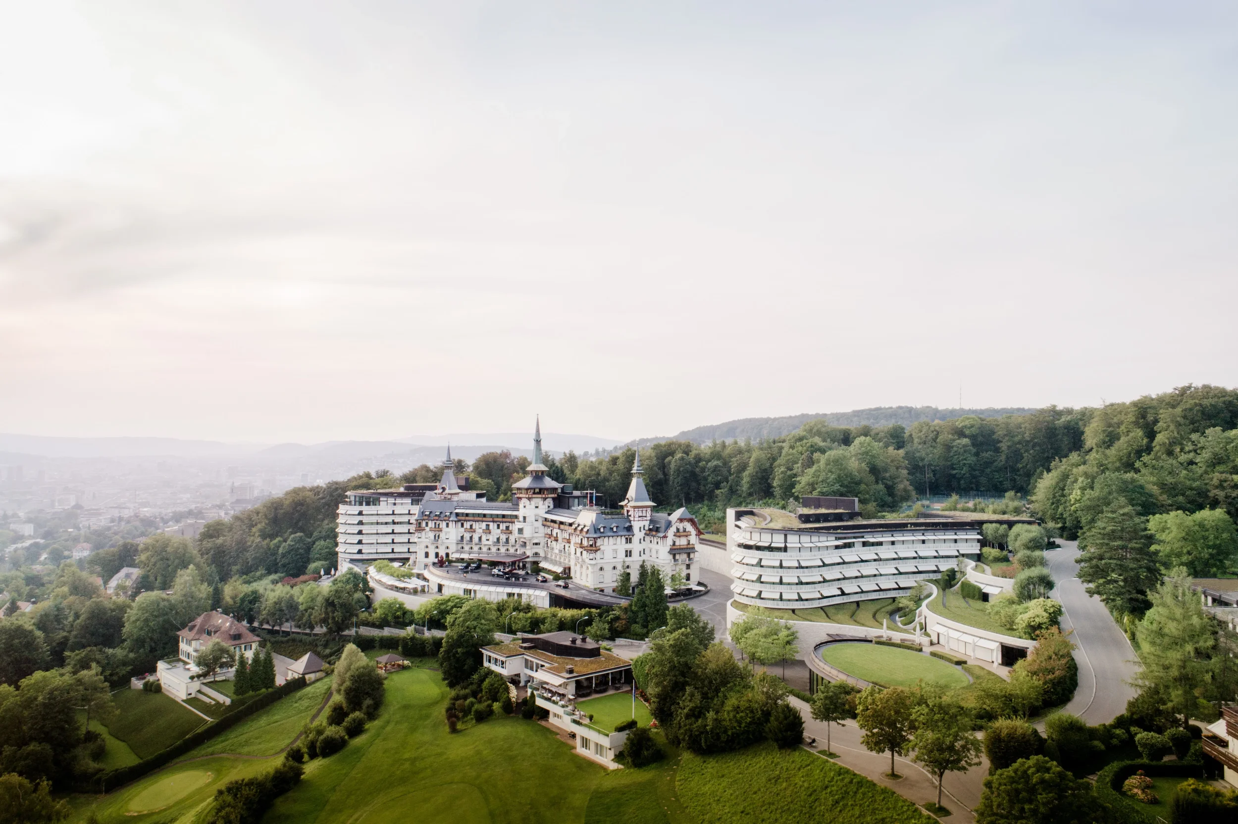 A large castle-like hotel with towers and turrets, surrounded by lush greenery and hills, with a modern parking structure nearby.